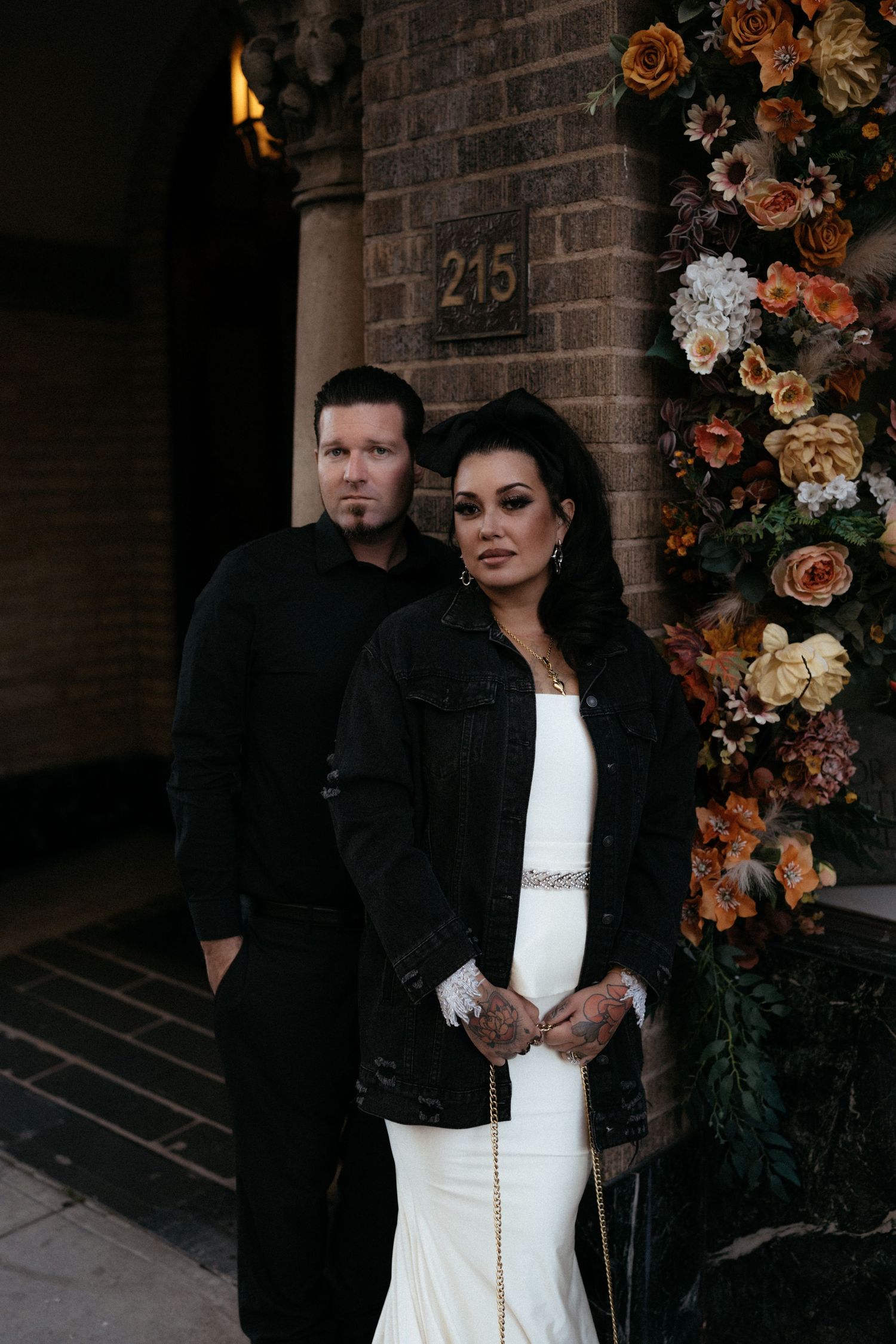 A couple poses together against a brick wall decorated with orange and white floral arrangements.