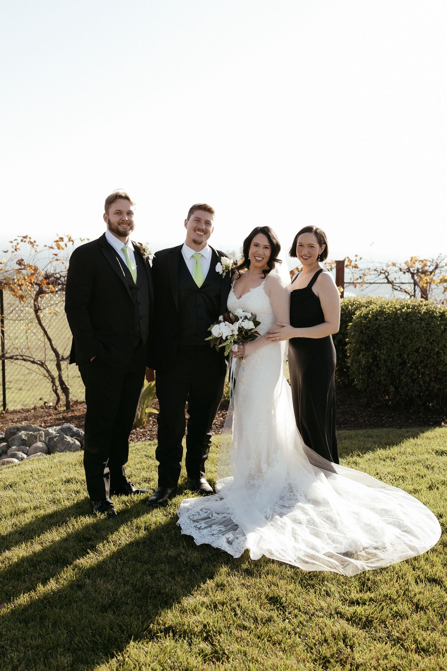 Wedding party poses together on grassy hillside during golden hour with flowing white dress and formal attire.
