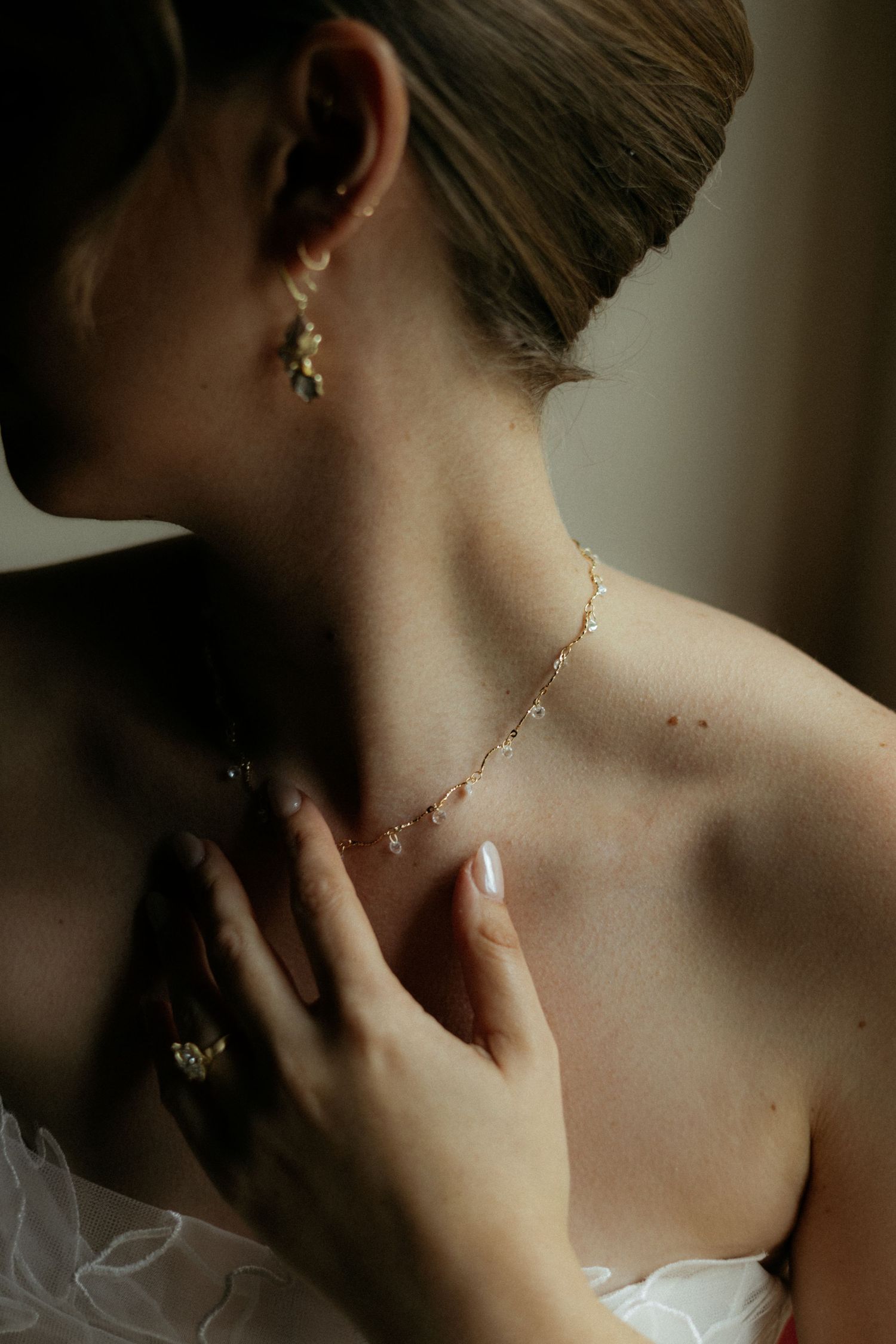 Bride wearing a delicate gold chain necklace and drop earrings, touching her collarbone with manicured hand.
