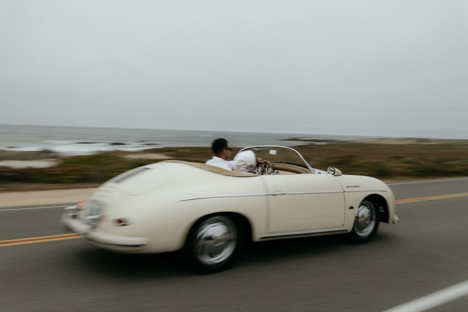 Couple driving a classic white Porsche 356 Speedster convertible along a coastal highway on an overcast day.