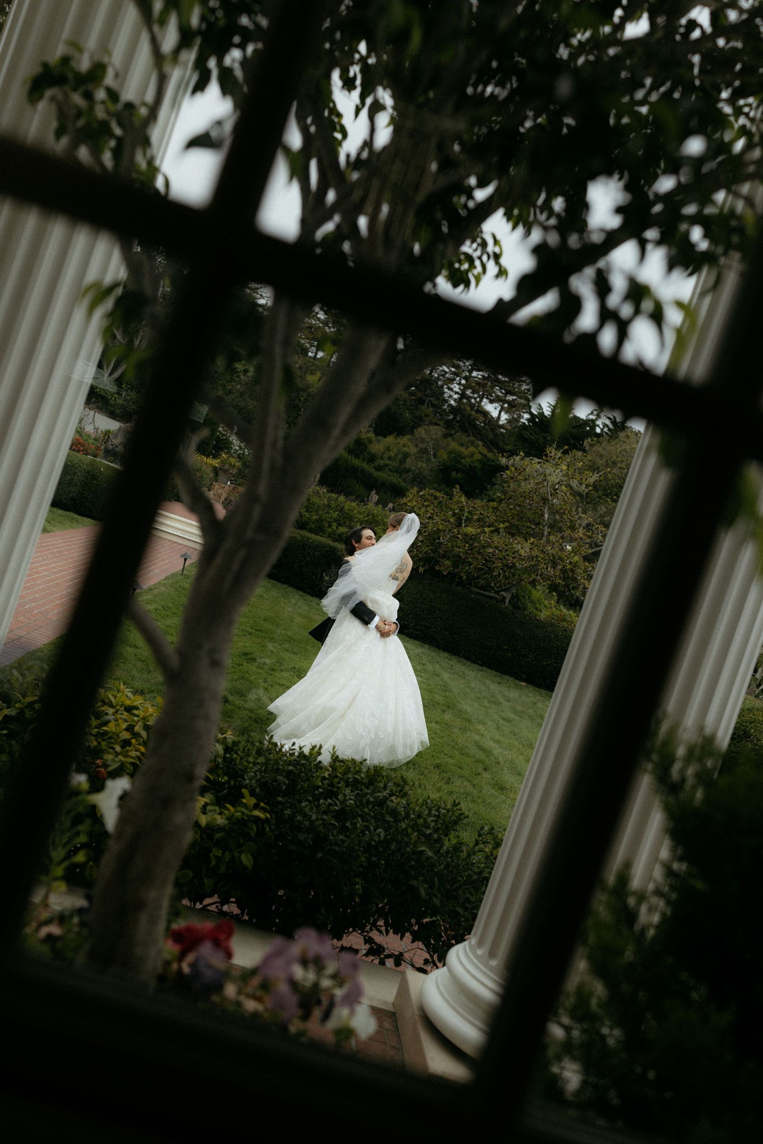 A bride and groom embrace in a lush garden, viewed through a white-framed window with columns.
