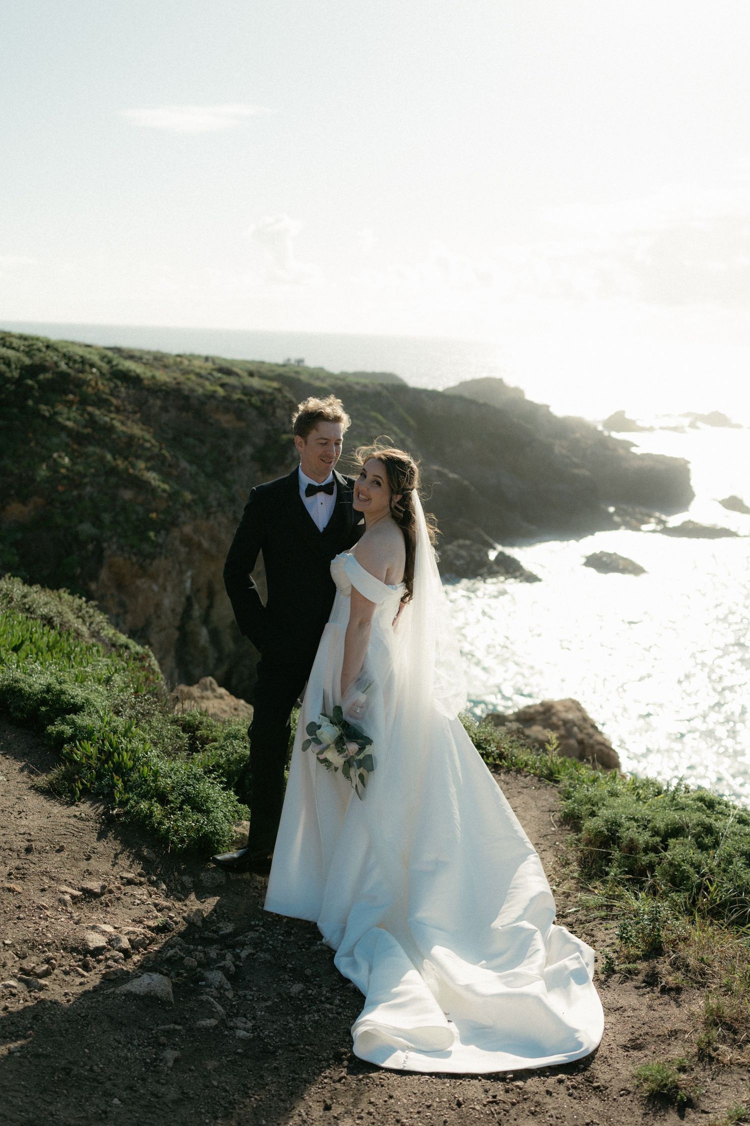 Elegant bride and groom pose on a rugged coastal cliff overlooking the ocean, bride holding a white bouquet in a flowing gown.