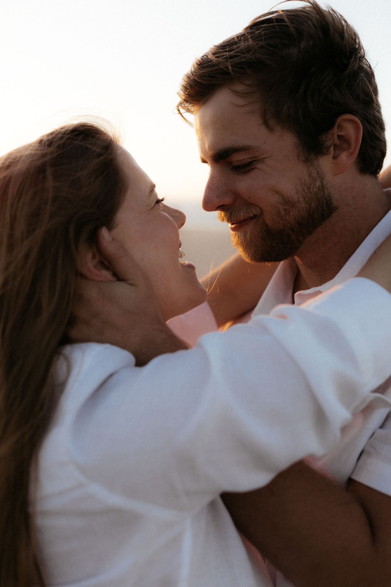 A romantic couple embracing and gazing into each other's eyes at sunset, both wearing white.