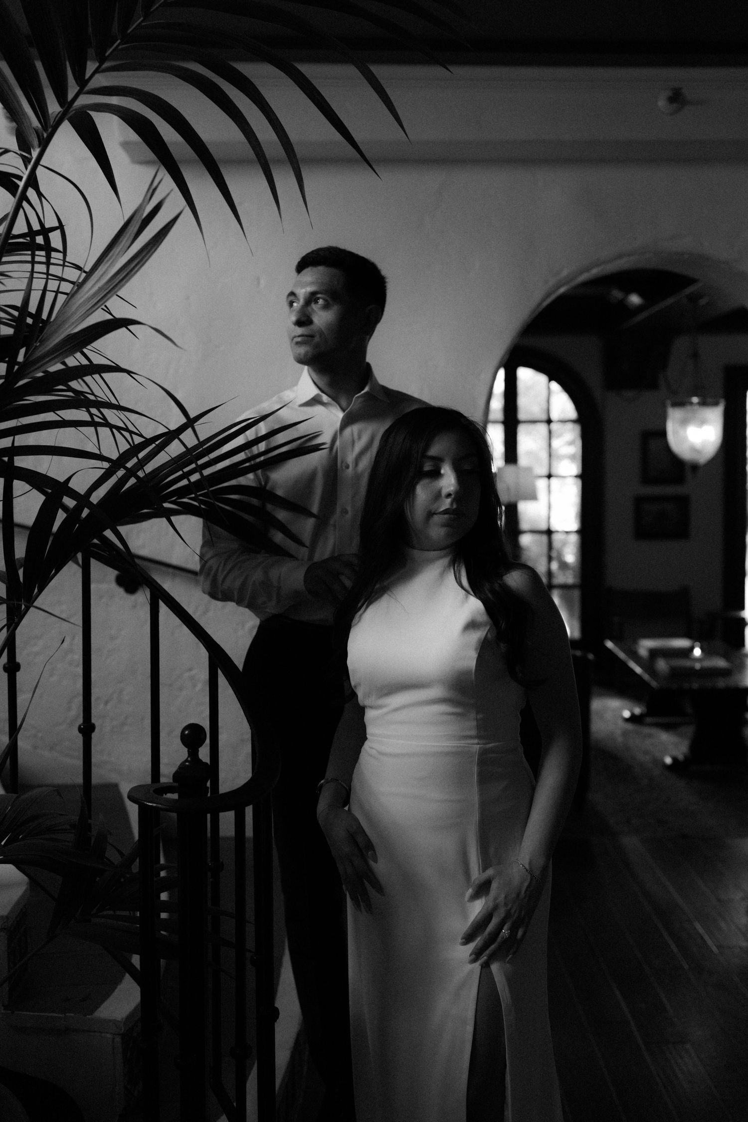 Black and white photo of an elegant couple posing indoors near a staircase and tropical plant in formal attire.
