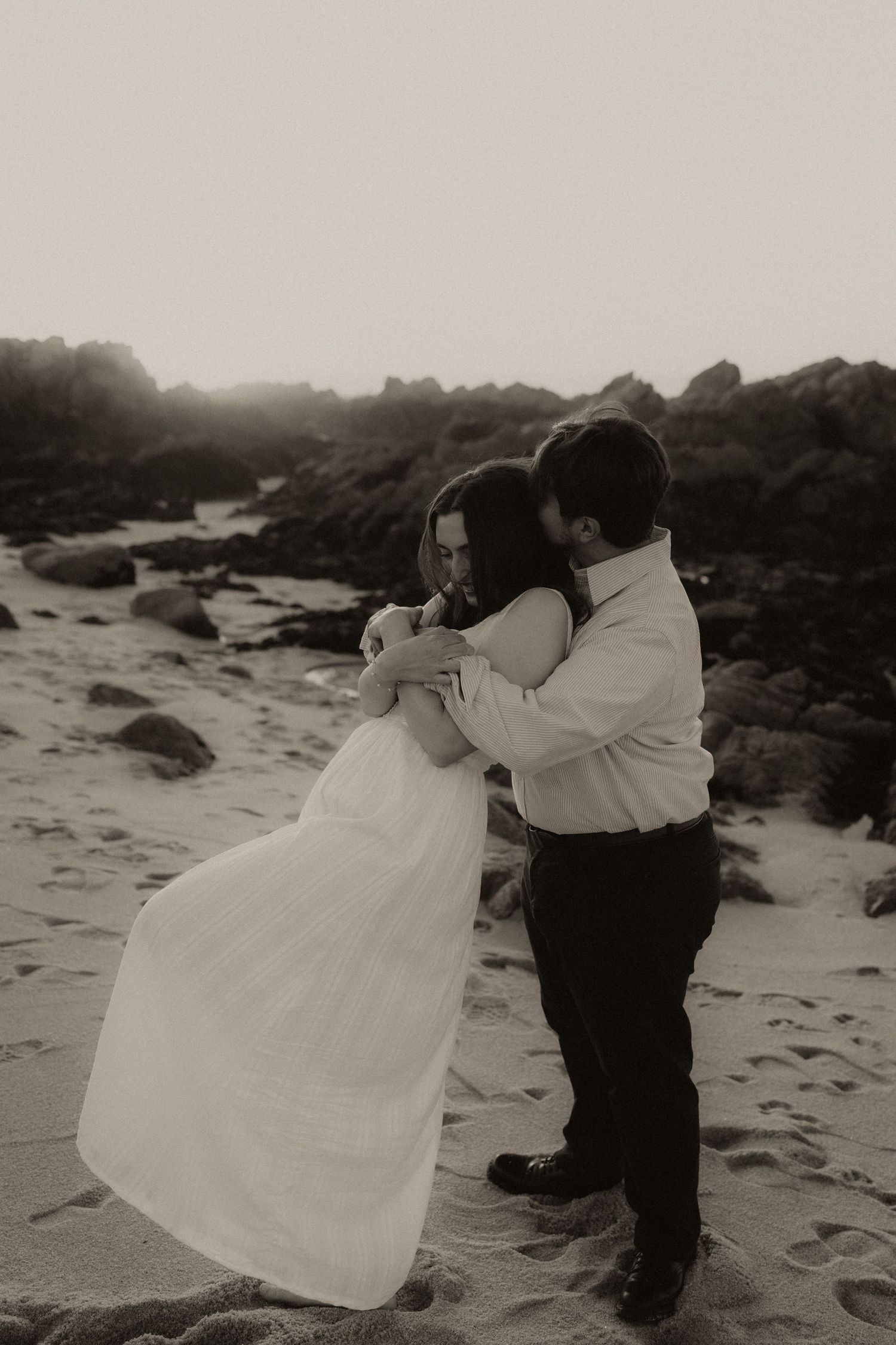 Black and white photo of a couple embracing on a sandy beach with rocky cliffs at sunset.