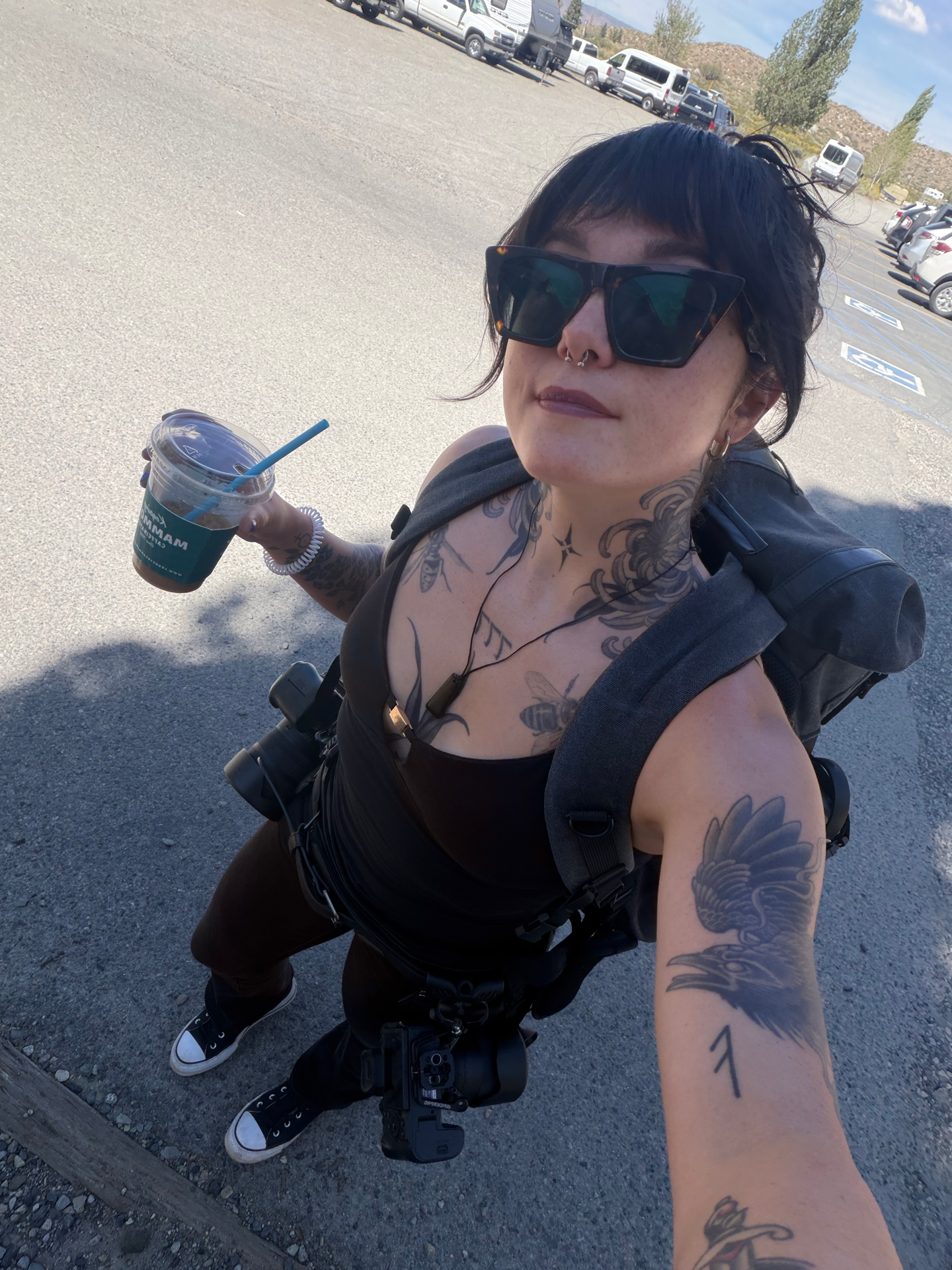 Tattooed woman in sunglasses holds iced coffee in a sunny parking lot, wearing a black outfit with a gray jacket.