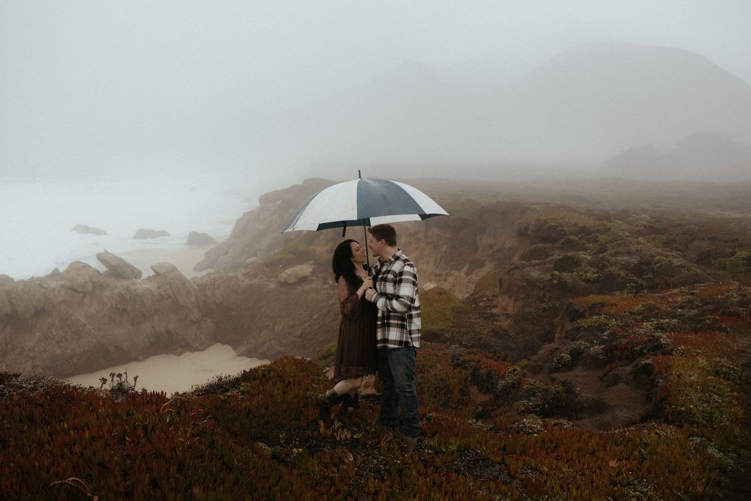Foggy coastal landscape with figures silhouetted against misty cliffs holding umbrellas.