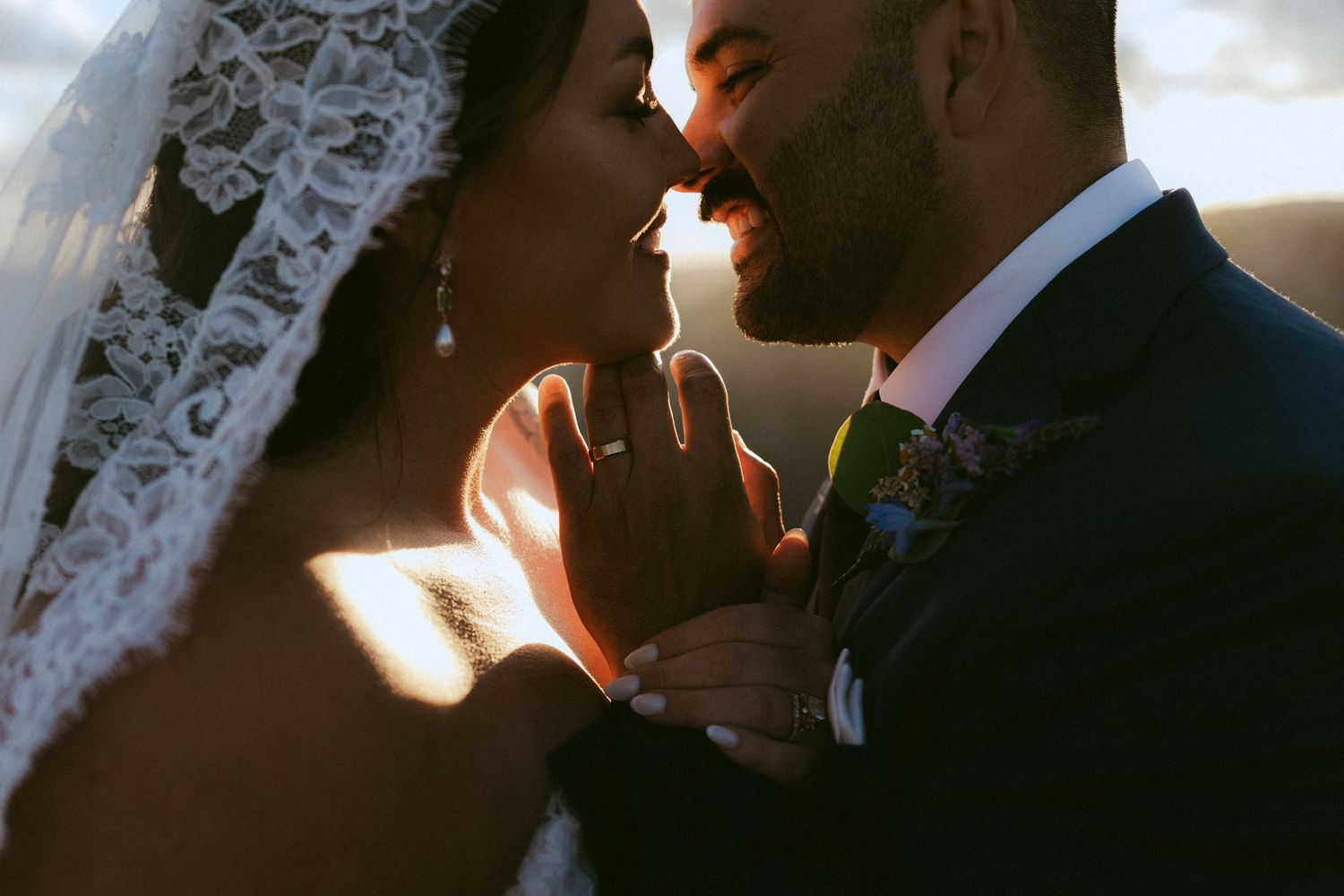 Bride and groom share an intimate golden hour moment, sun rays illuminating lace veil during wedding ceremony.