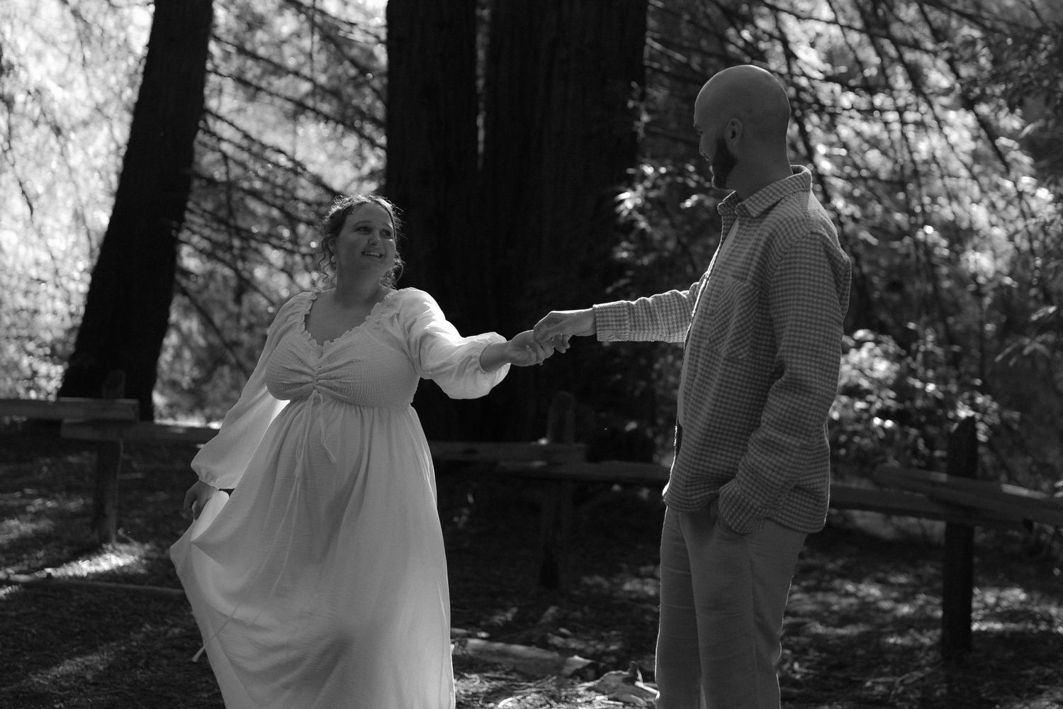 A couple dances together in a forest setting with dappled sunlight filtering through the trees.