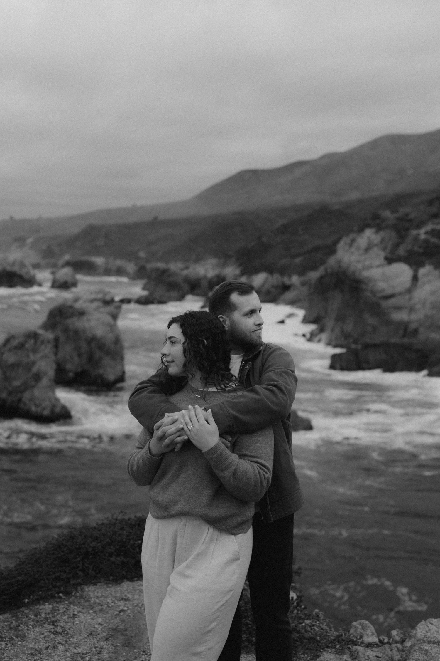 A couple embraces while standing on a rugged coastal cliff with dramatic ocean waves and rock formations in the background.