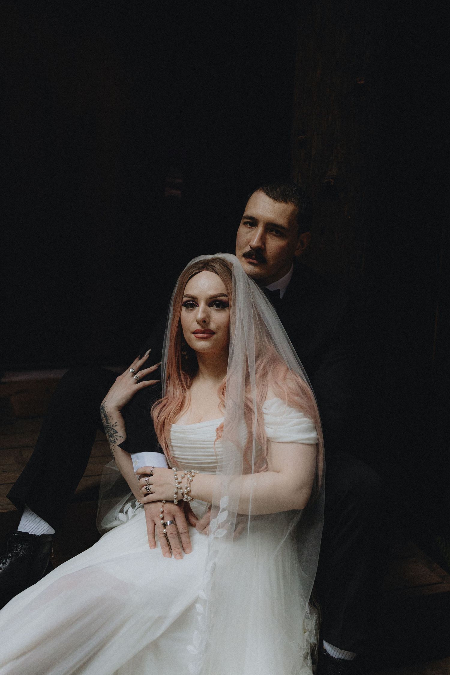 Moody wedding portrait showing couple in traditional attire with bride in white dress and veil against dark background.