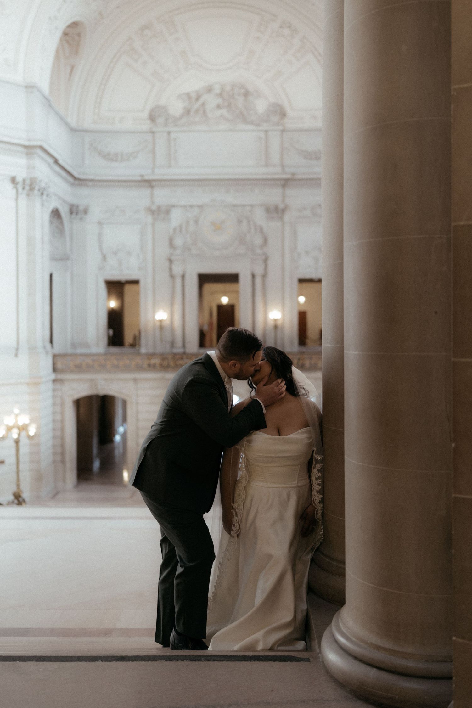 Romantic wedding couple shares intimate moments between elegant marble columns in a grand historic building.
