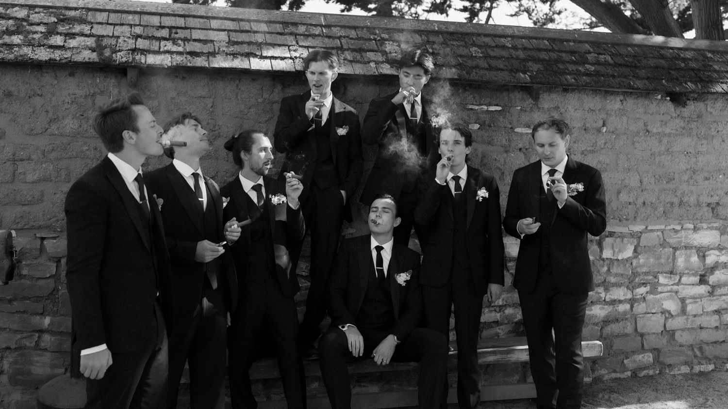 A black and white panoramic photograph shows a large group of formally dressed men standing in a line against a brick wall.
