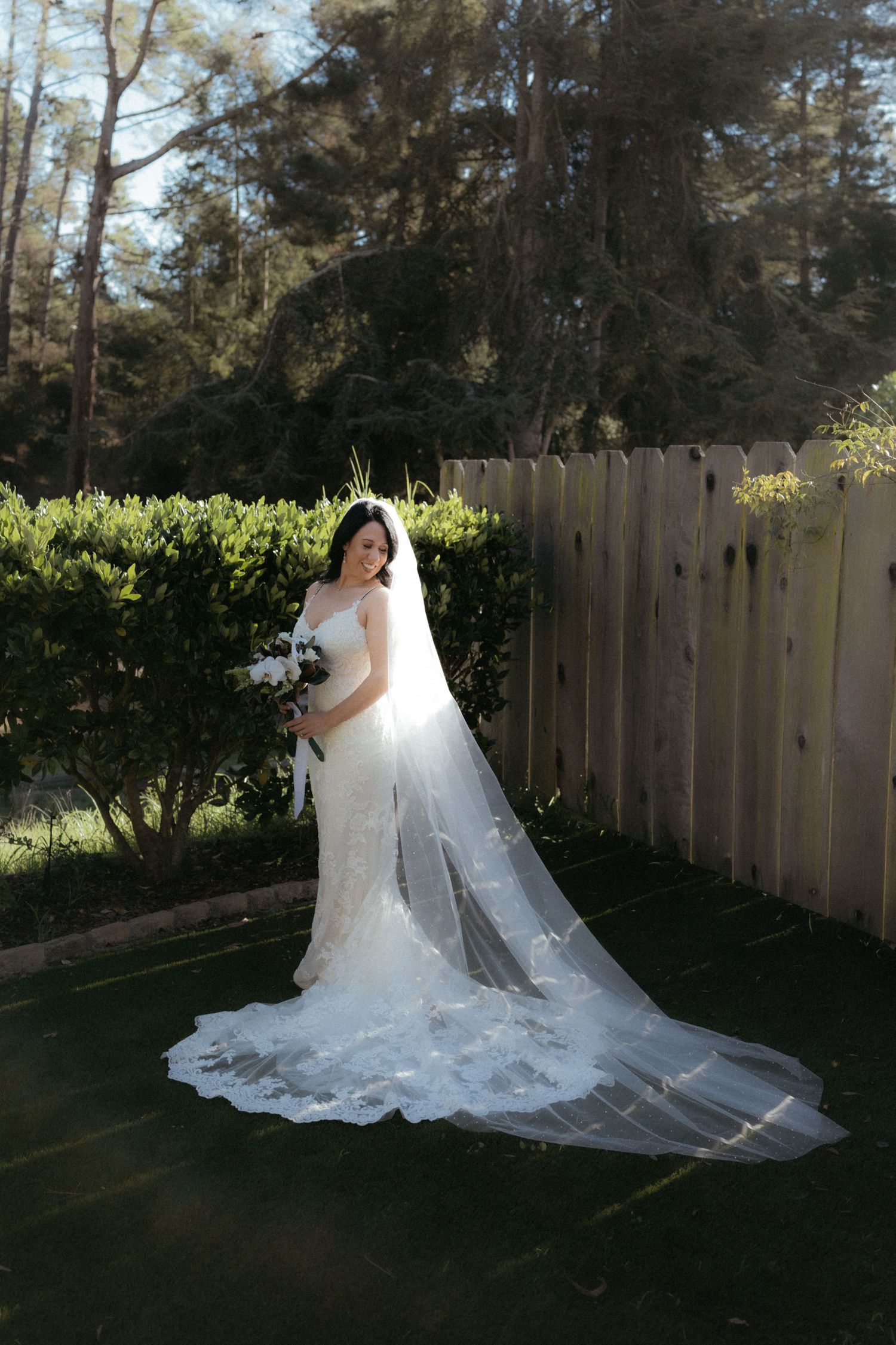 A flowing white wedding dress with a cathedral-length veil creates an elegant silhouette against a wooden fence backdrop.