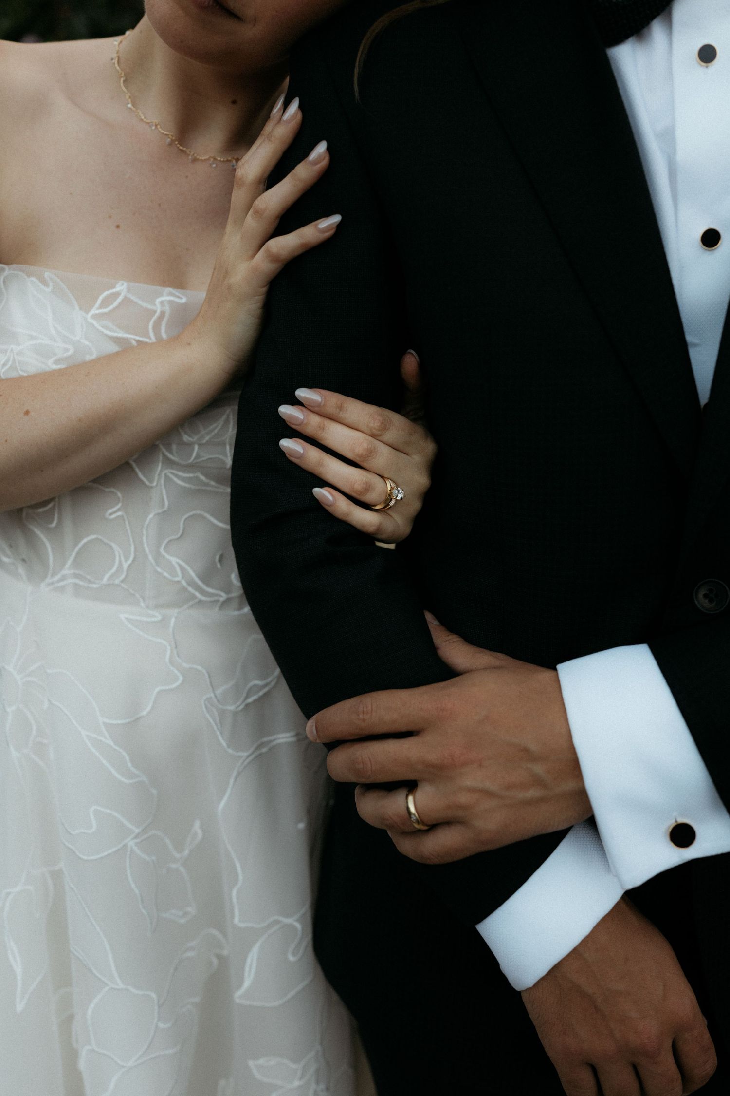 Close-up of bride and groom showing wedding rings, bride in floral strapless gown holding grooms arm.