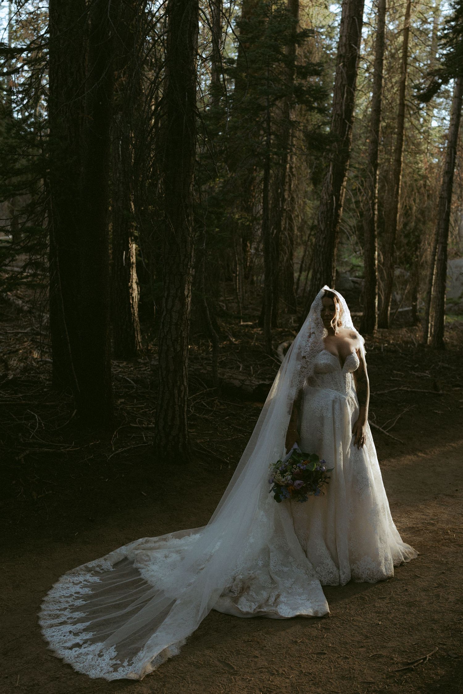 Bride in white gown and long lace veil holding bouquet in a dark forest setting.