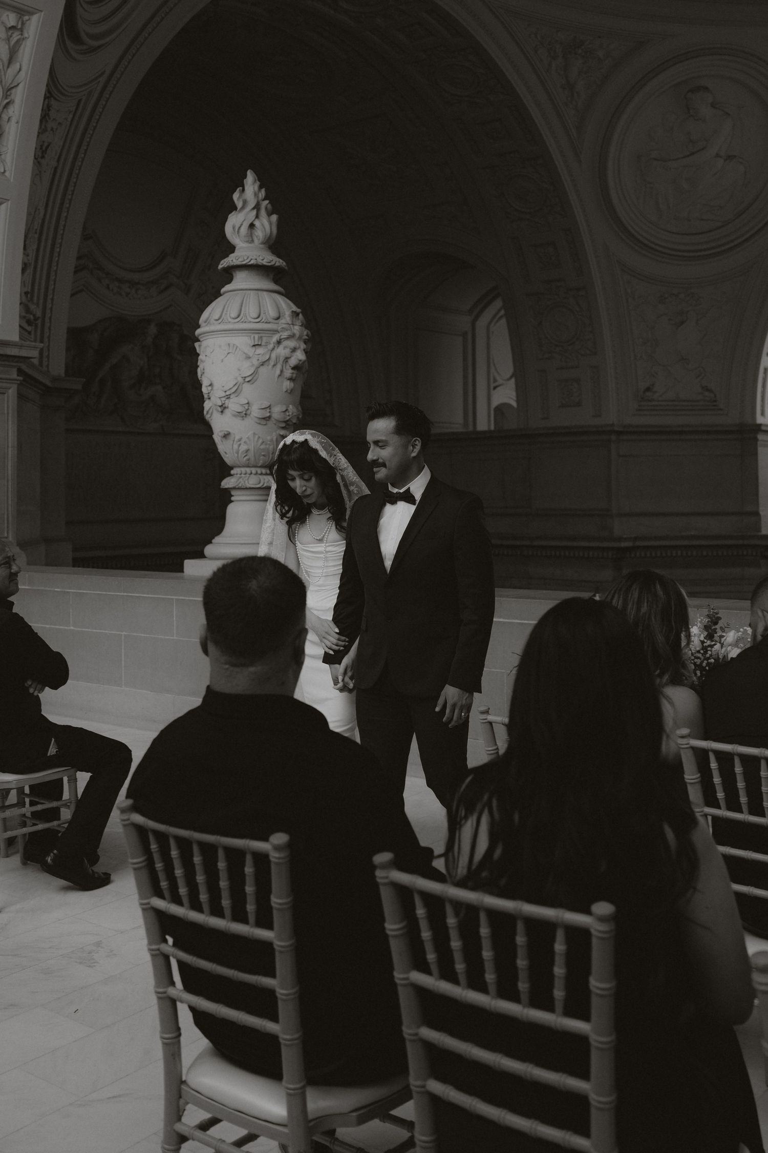 Wedding guests seated in an ornate hall with classical statues and architectural details.