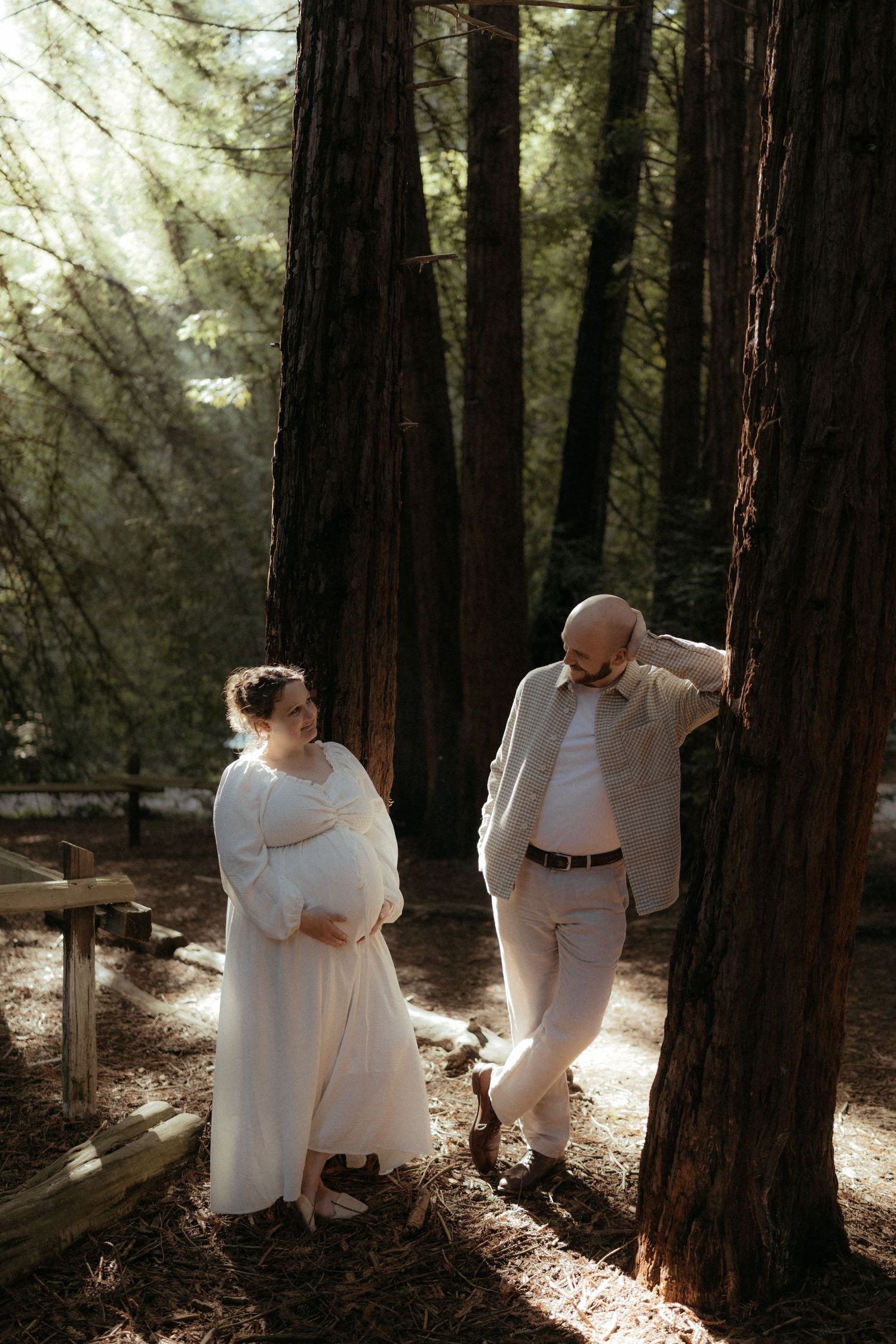 A couple in matching white outfits shares an intimate moment among majestic redwood trees.