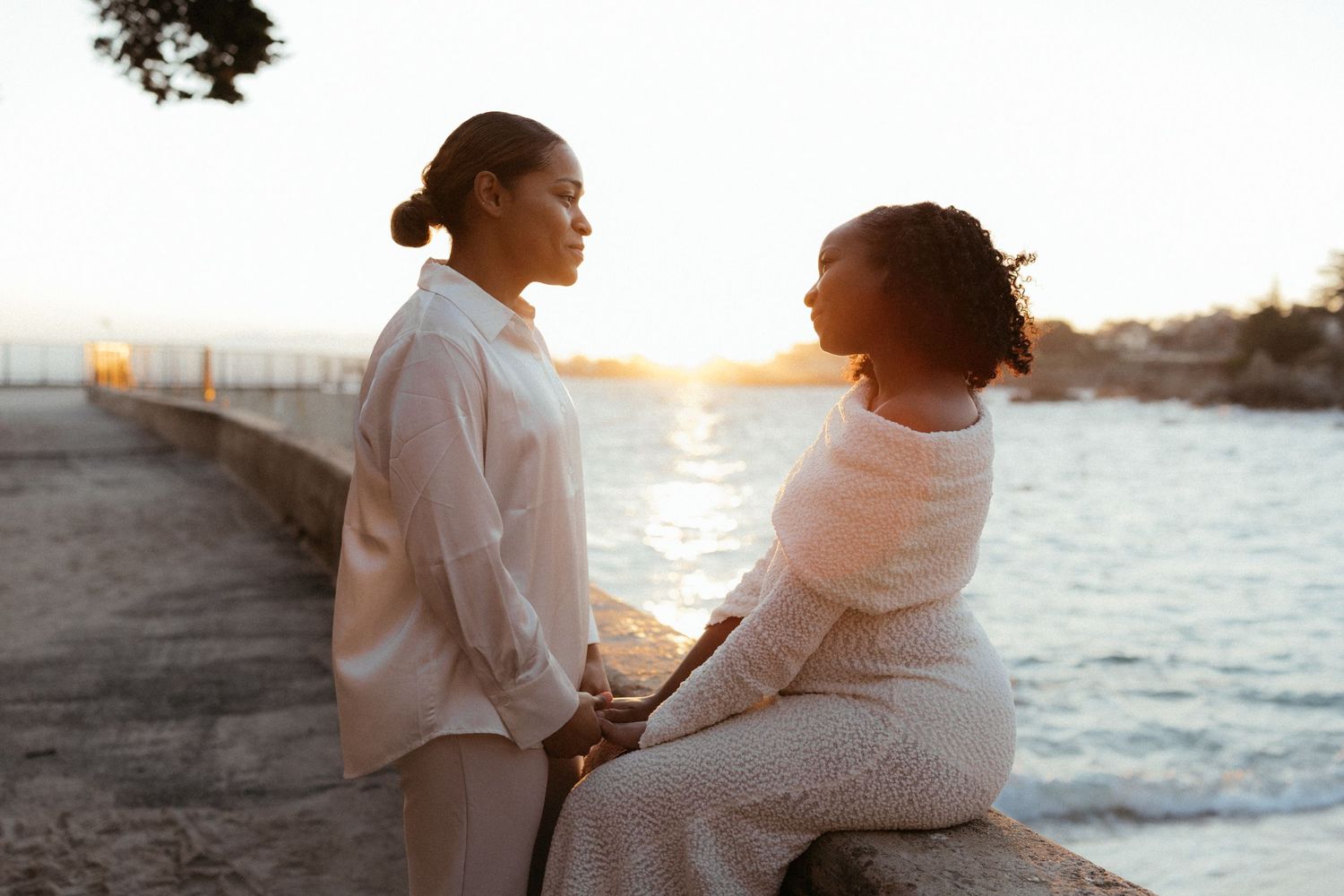 Two people in white outfits share a tender moment while sitting by the ocean at sunset.