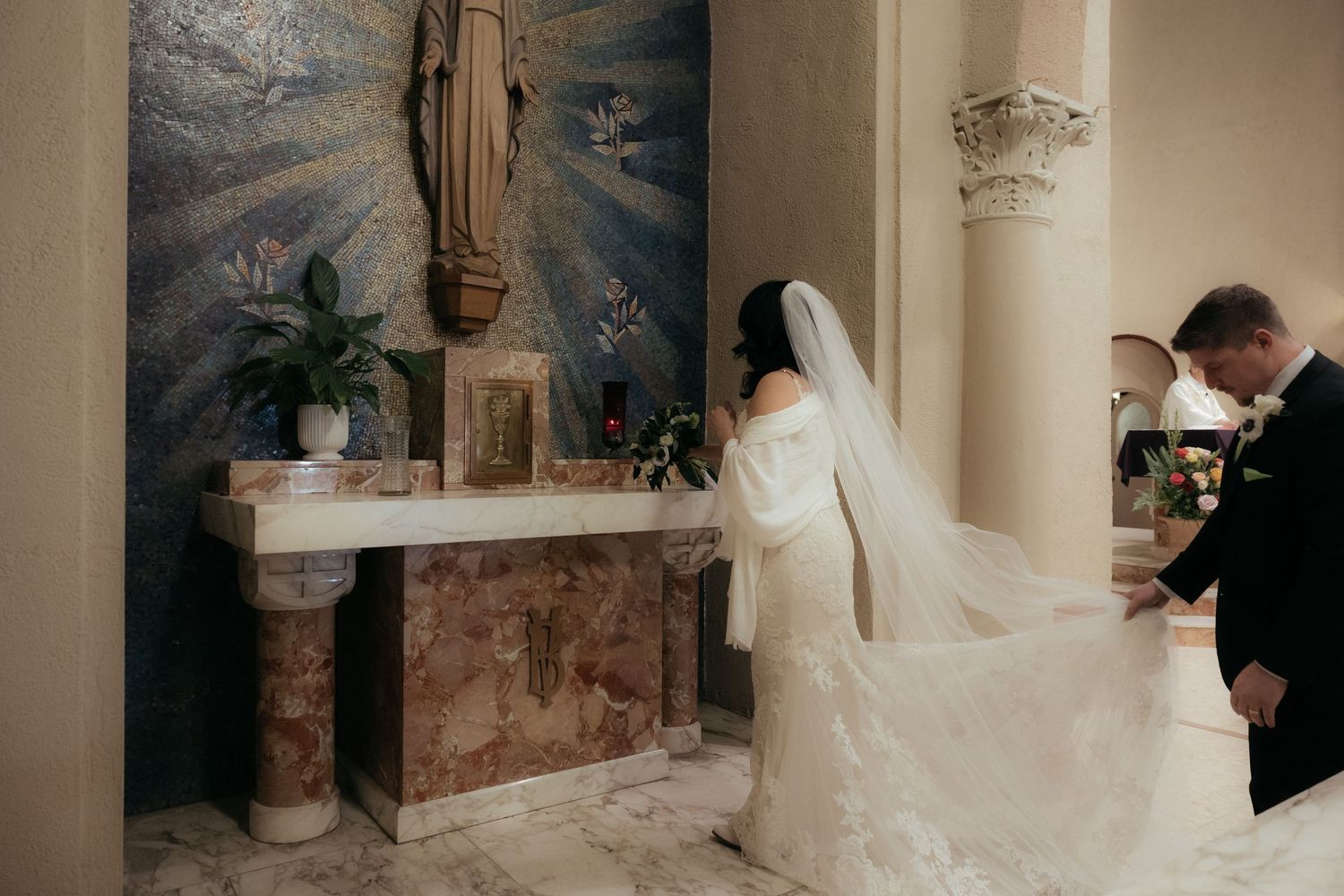 A bride in a white wedding gown and veil kneels at a church altar with decorative marble columns and religious artwork.