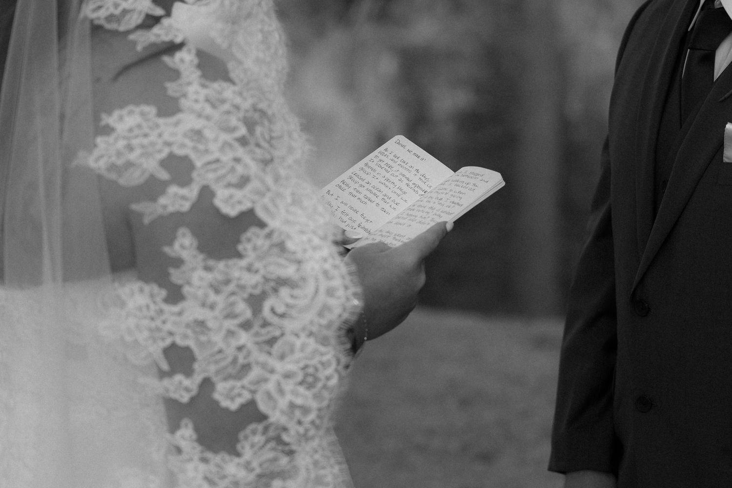 Close-up black and white photo of groom reading wedding vows from a small book beside bride in lace gown.