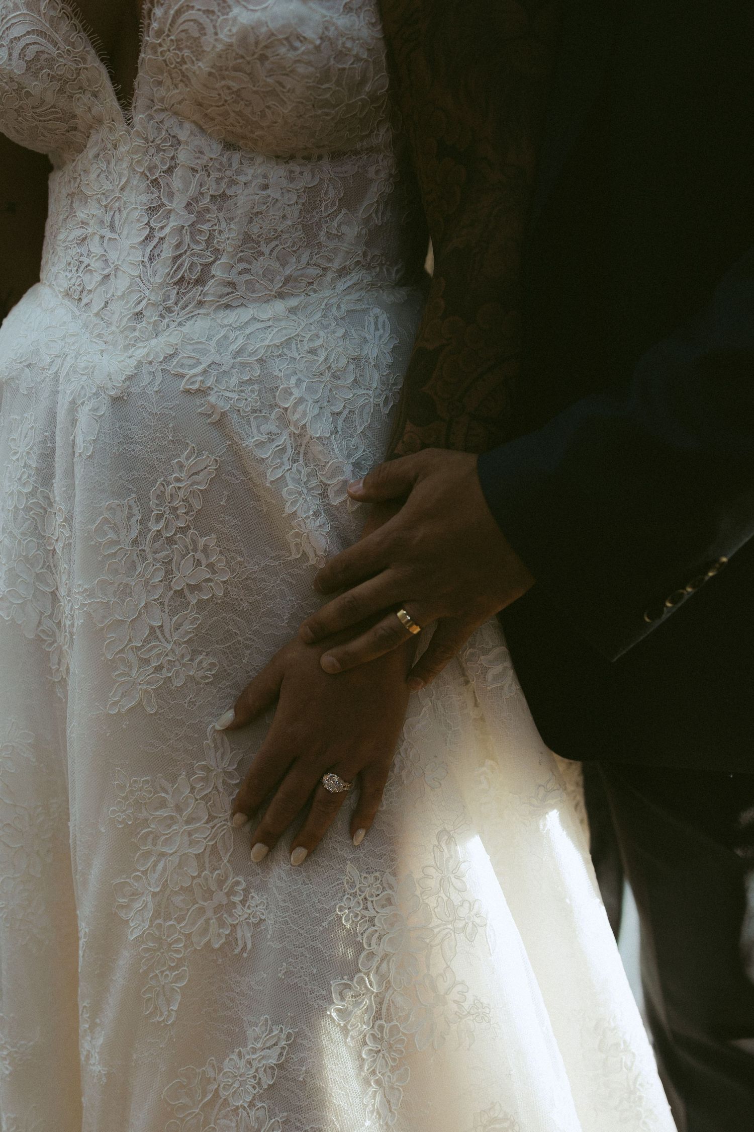 Close-up of bride and groom holding hands, showcasing wedding rings on lace bridal gown.