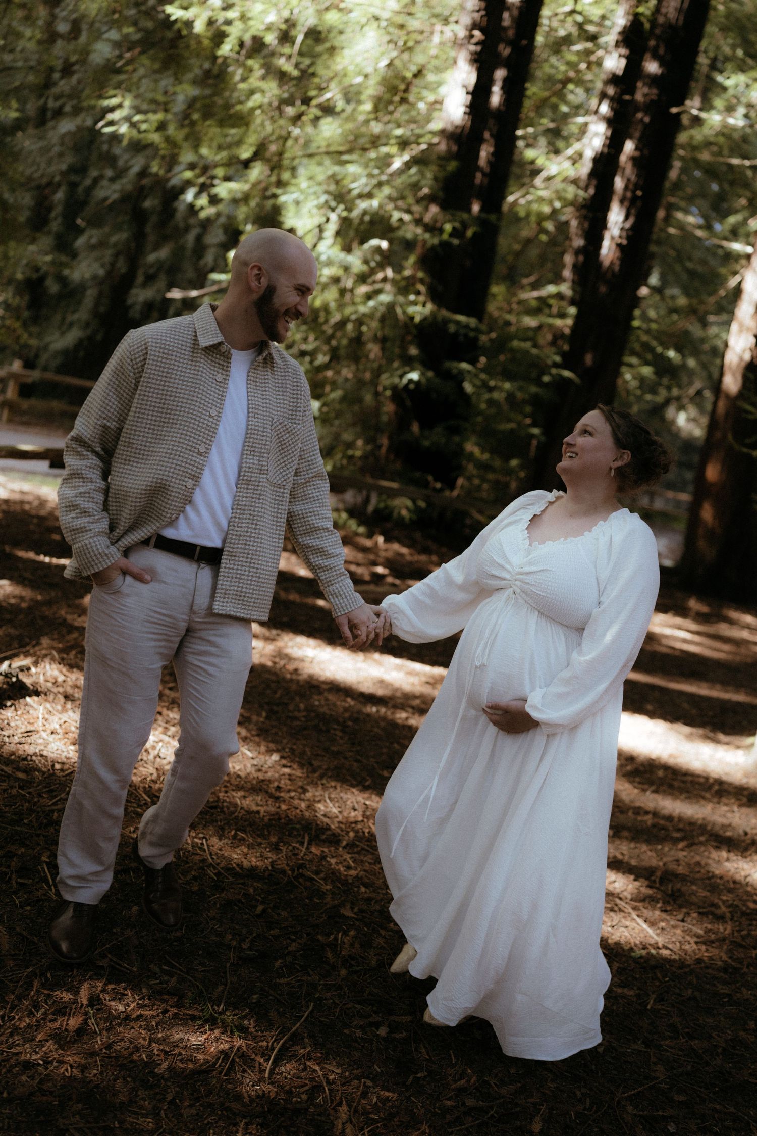 A couple shares intimate moments in a sunlit forest setting wearing coordinated white and beige outfits.