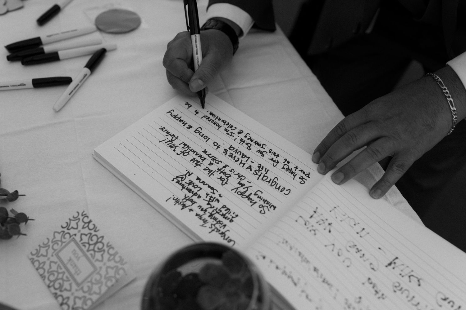 Close-up view of someone writing in a guest book with pens scattered on the table.