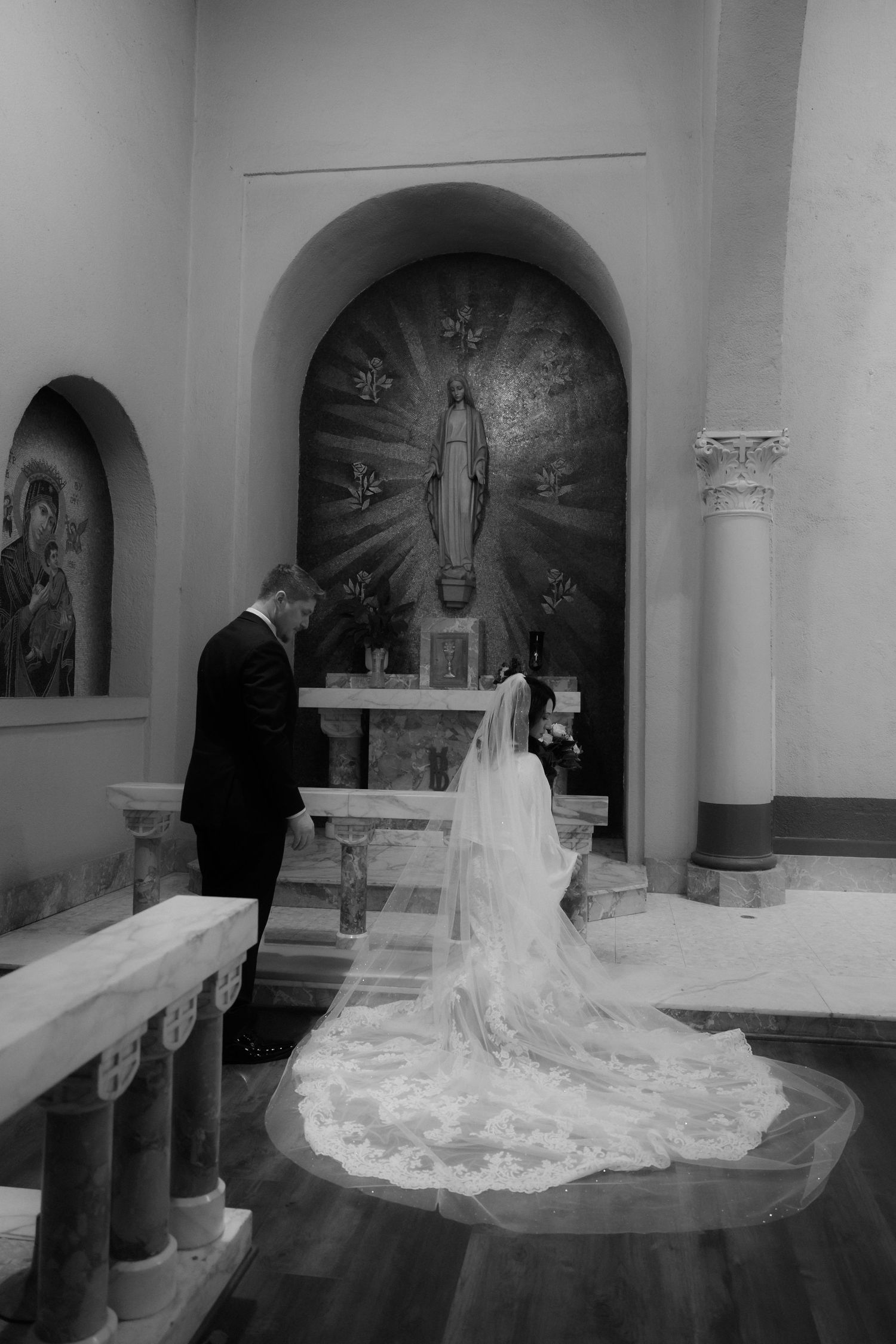 A bride in a flowing white gown with a long cathedral veil kneels at the altar in a church sanctuary.