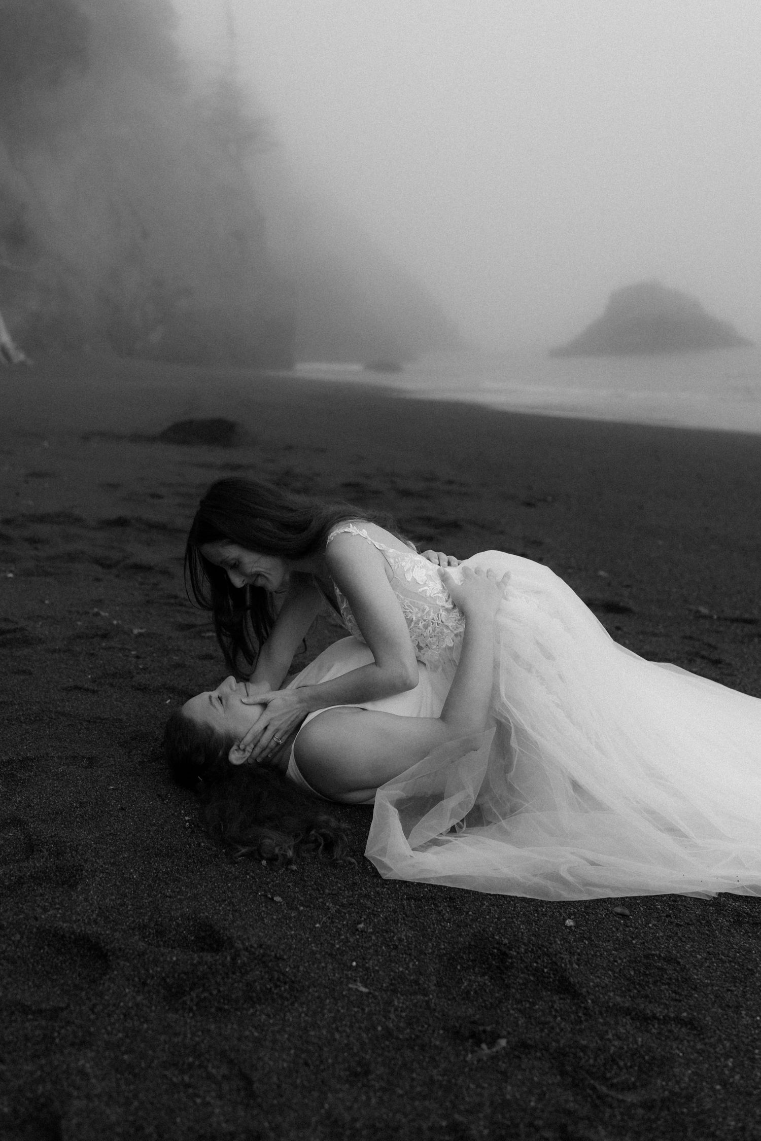 A figure in a white wedding dress lies on a dark volcanic beach in a moody, atmospheric black and white photo.