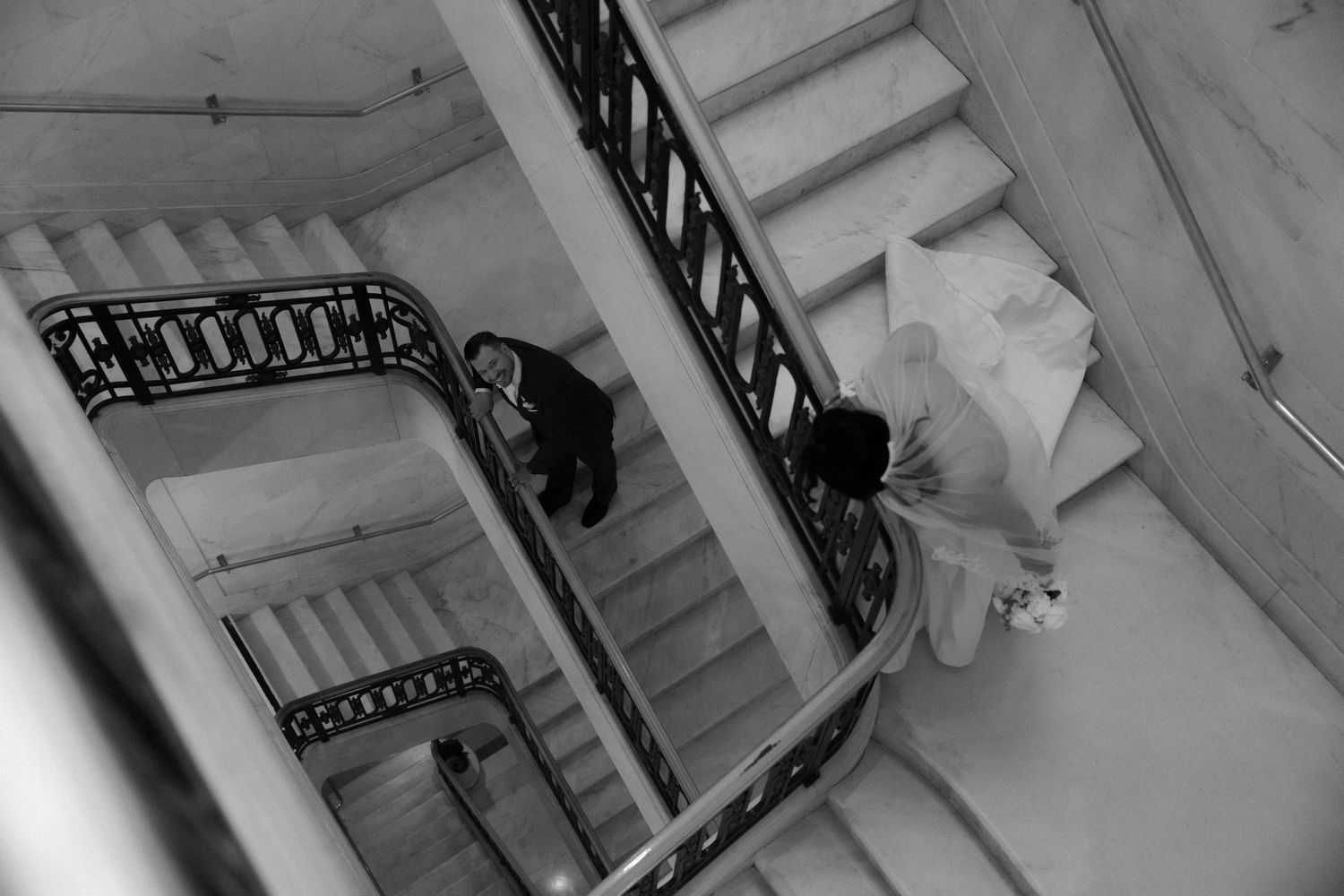 A black and white aerial view of a bride descending an ornate spiral staircase with wrought iron railings.