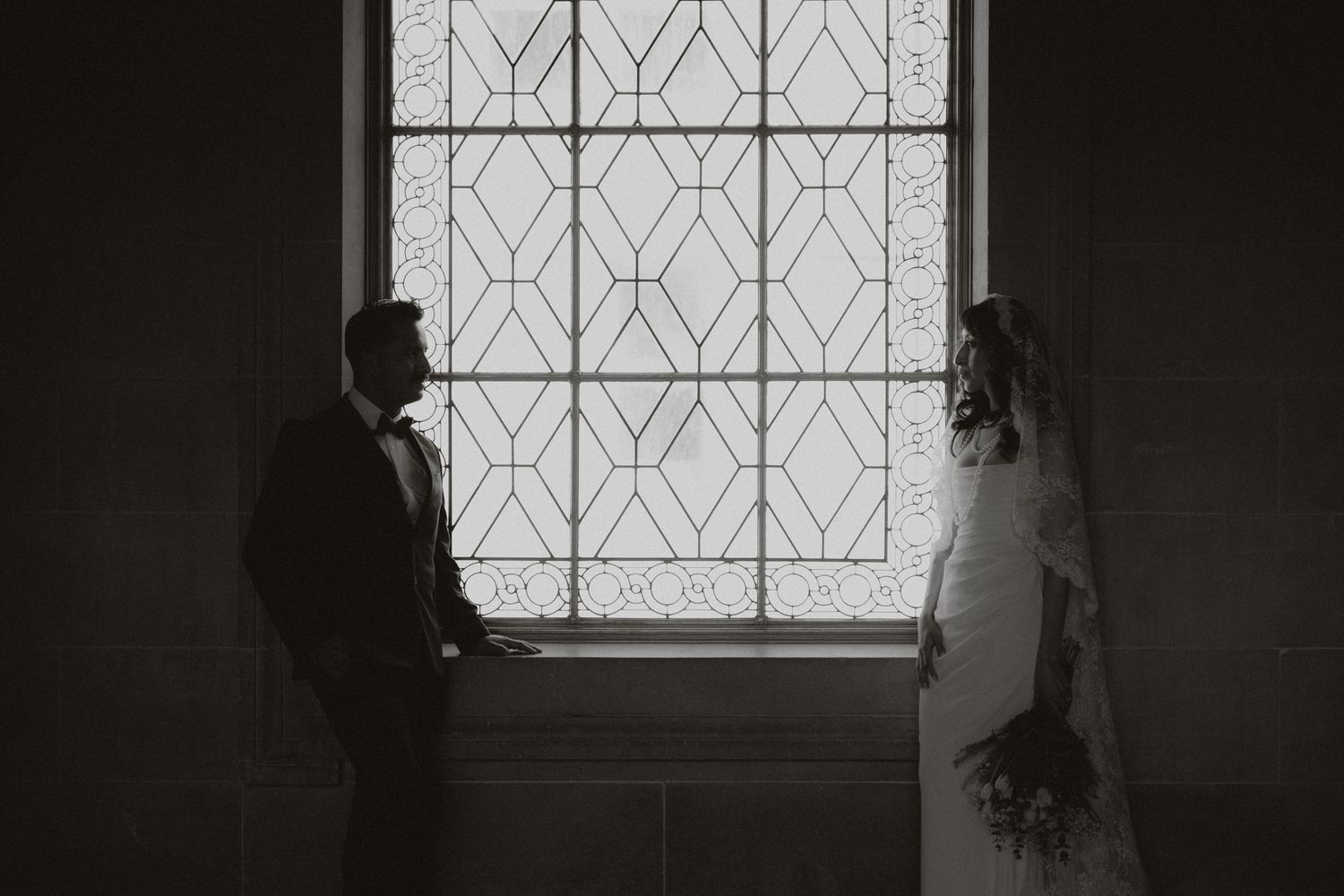 A dramatic black and white photo of newlyweds standing by a geometric window.