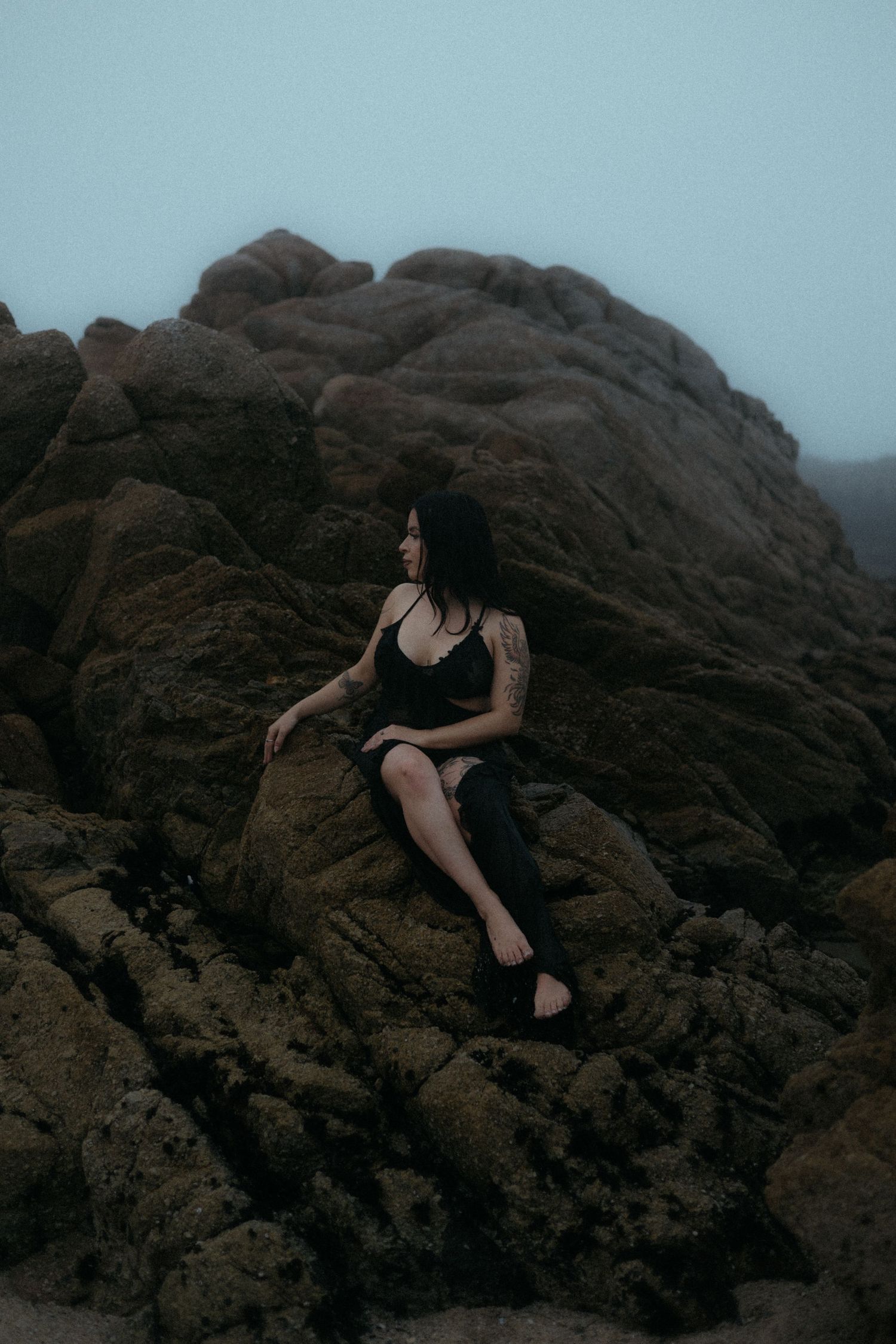 Figure in black reclines on dramatic rocky coastline during misty evening.