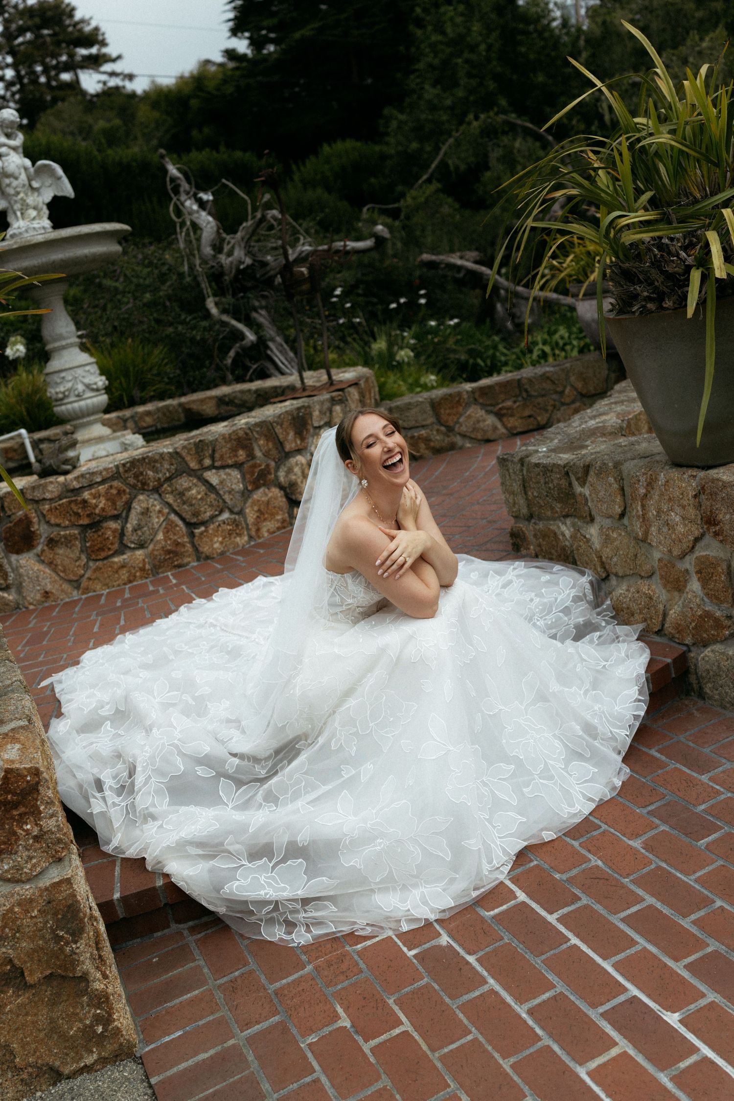 Joyful bride in lace wedding gown and veil sitting on brick steps laughing in garden setting.
