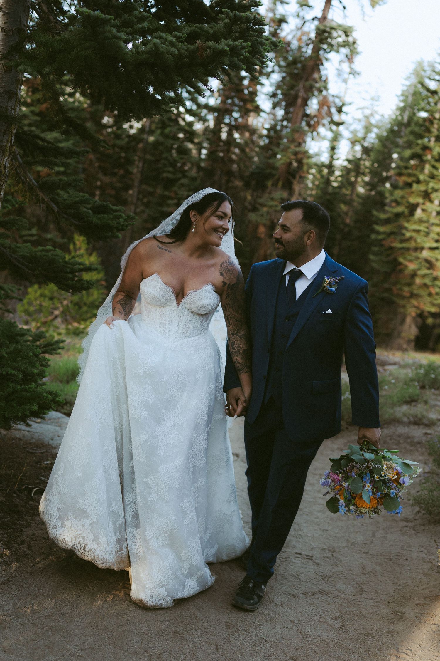 Bride and groom walk hand-in-hand through a forest path, she in lace gown with veil, he in navy suit holding bouquet.