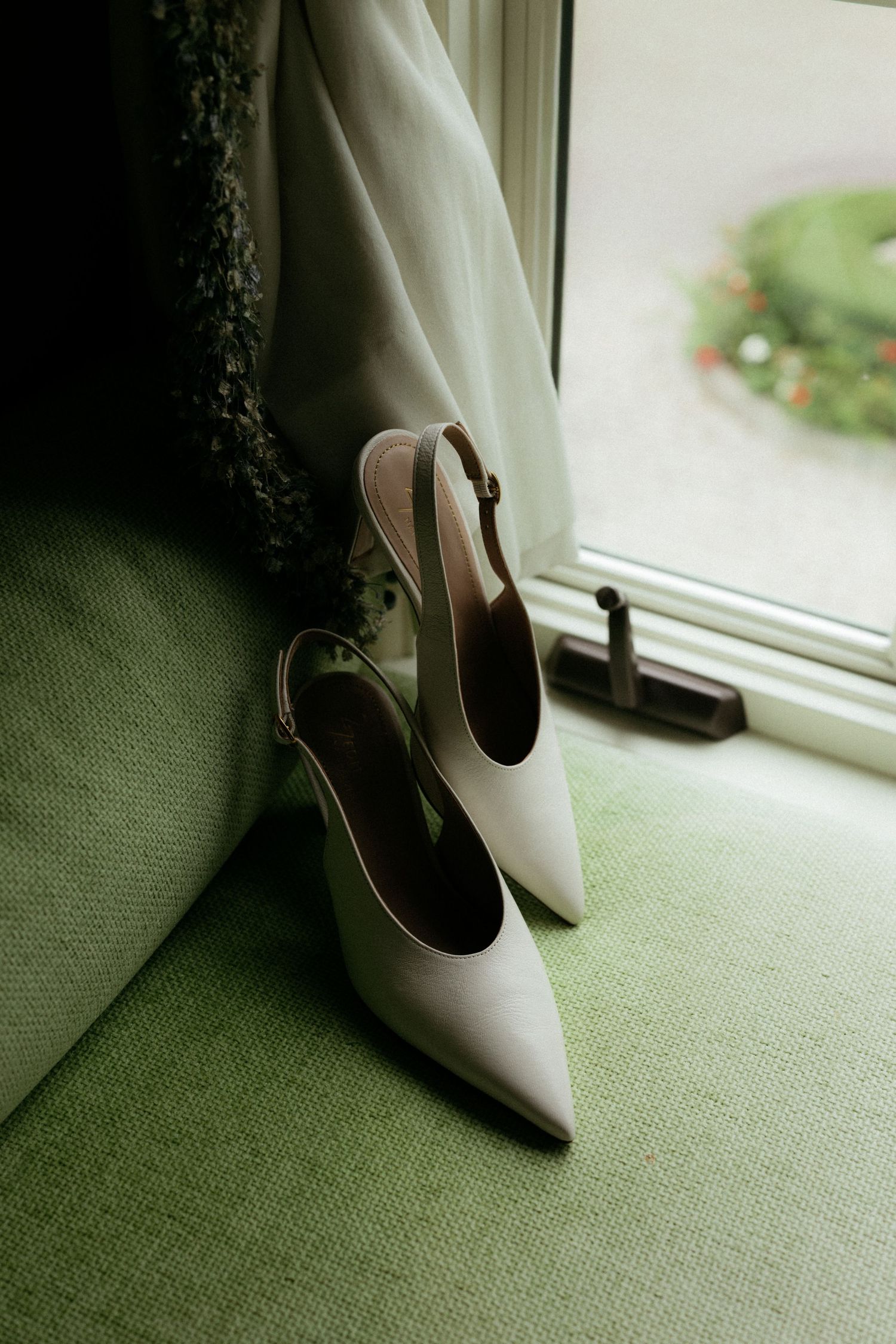 Elegant white slingback pointed-toe bridal heels rest on a green chair beside a window on wedding day.