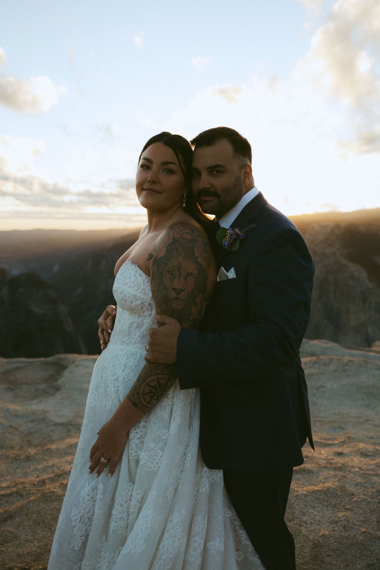 Bride and groom embrace at golden hour on a rocky mountain overlook, bride wearing lace gown with sleeve tattoo.