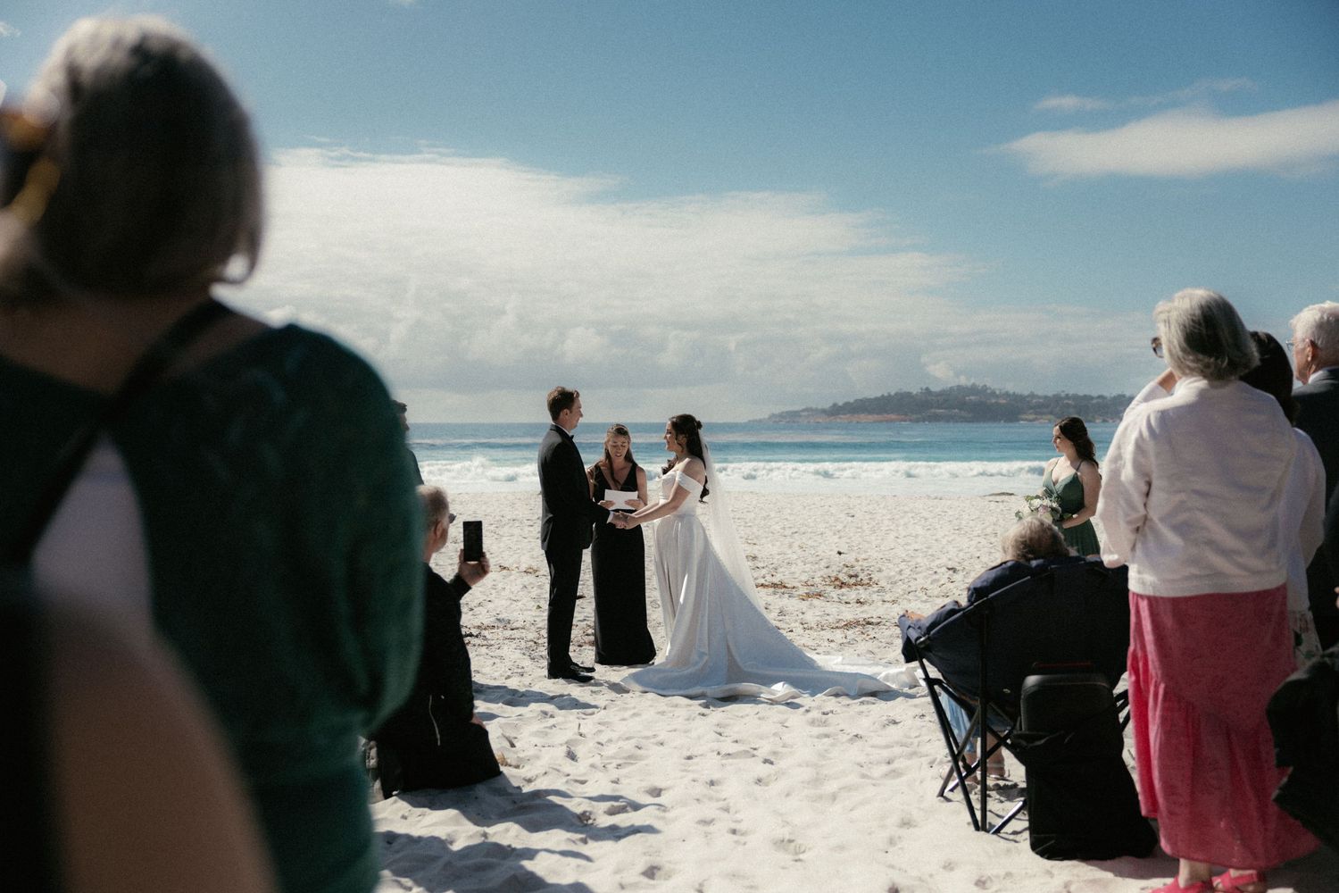 Beach wedding ceremony with bride in white gown and groom, officiant in black, guests seated on white sand with ocean backdrop.