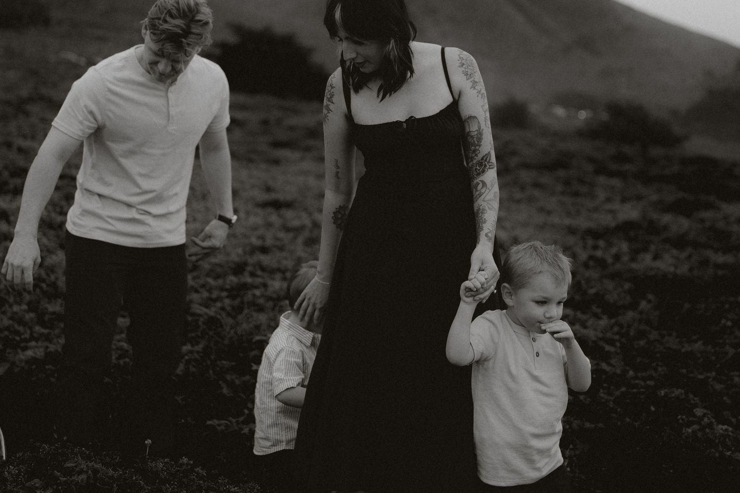 Black and white family photo of parents and toddler walking in a green meadow, mother in black dress with tattoos.