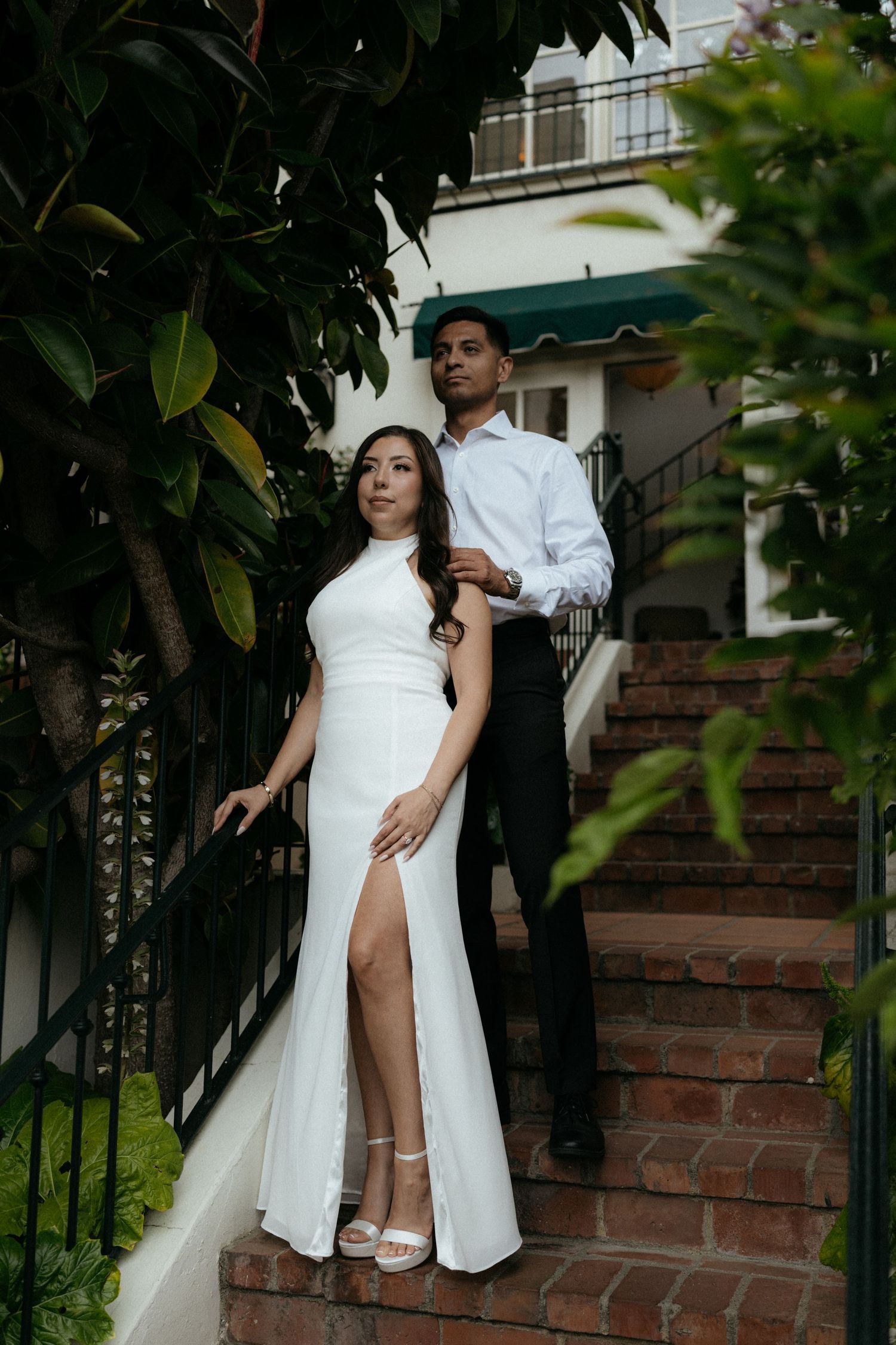 Couple posing on brick steps surrounded by lush greenery, woman in white slit gown, man in white shirt.