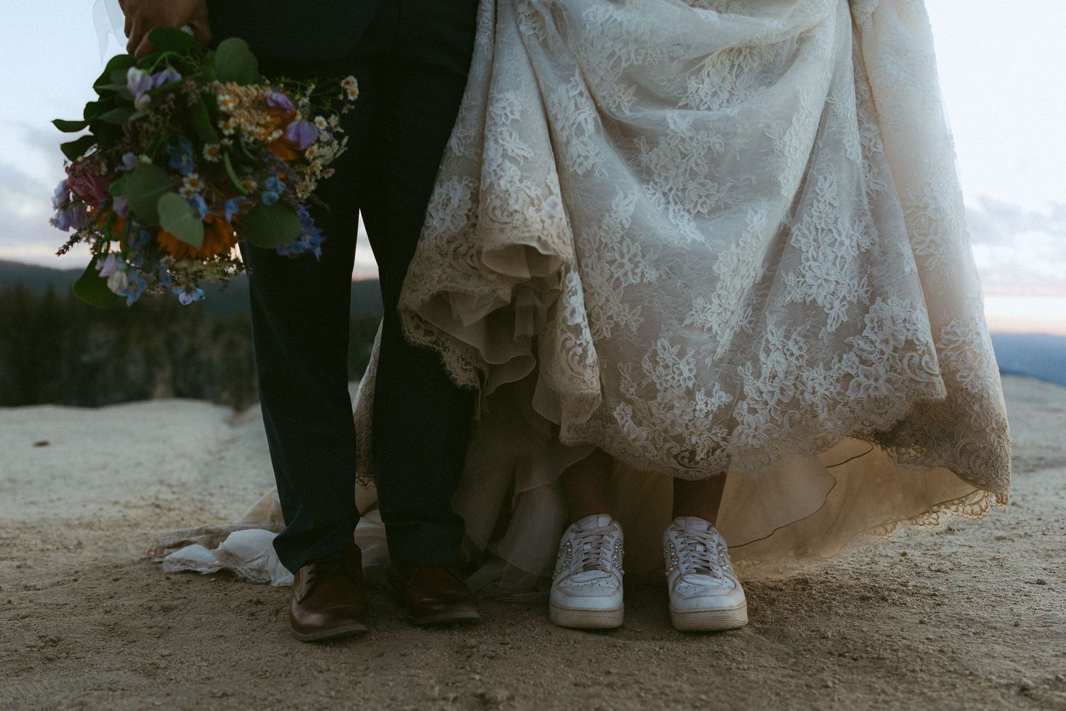 Bride in lace wedding gown with white sneakers and groom in dark suit holding colorful bouquet standing on rocky ground.
