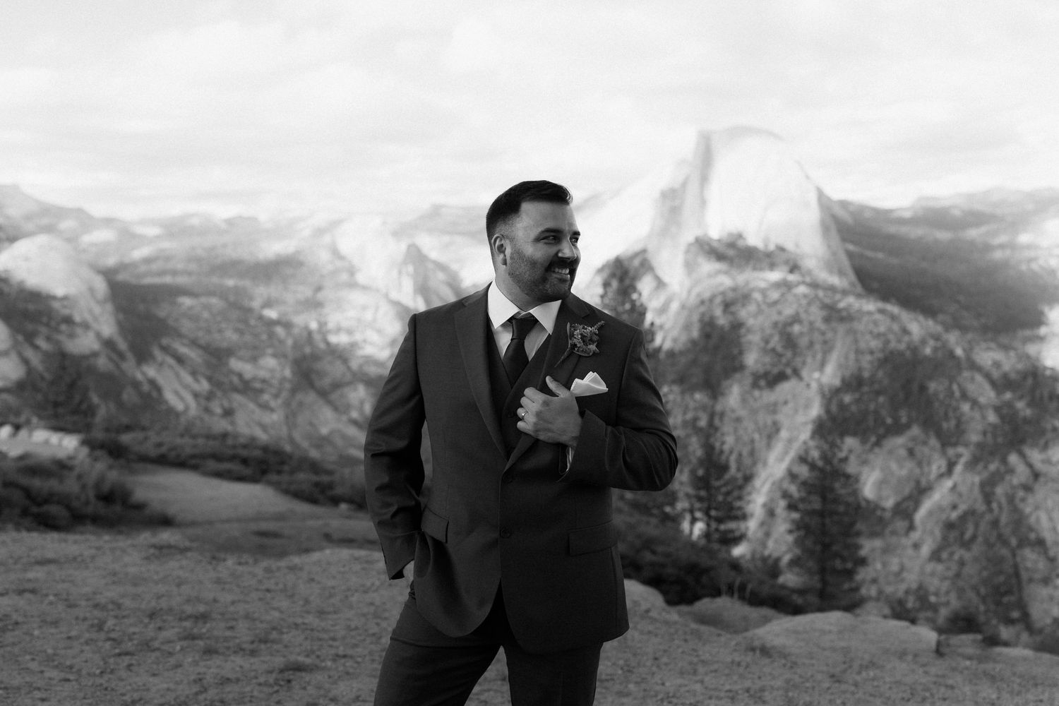 Groom in dark suit with boutonniere posing confidently before Half Dome at Yosemite in black and white.