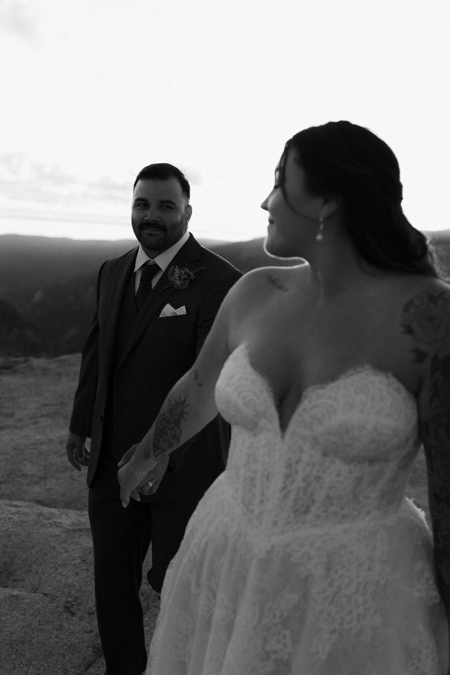 Black and white photo of bride and groom holding hands on mountain overlook at sunset.