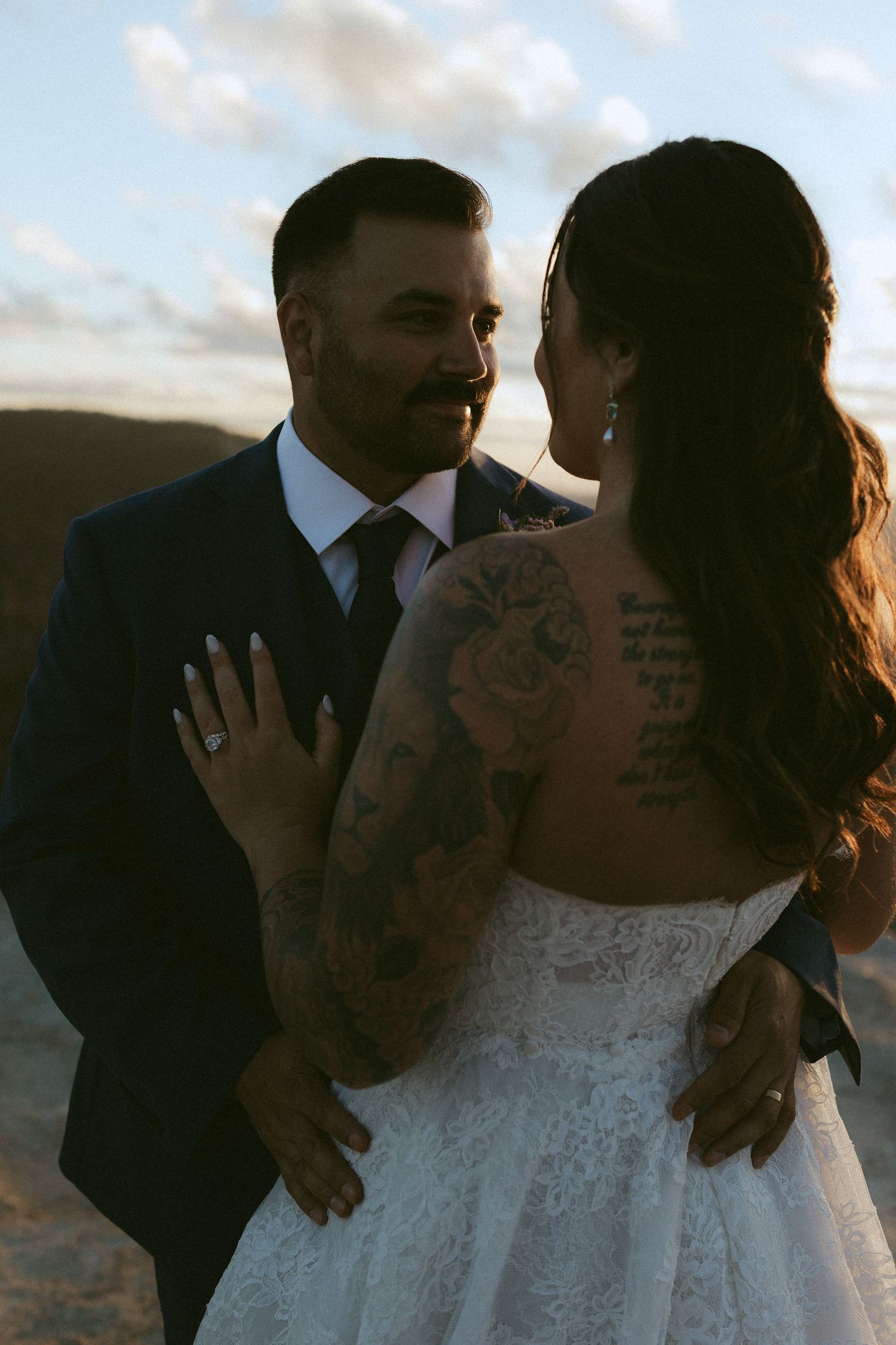 Tattooed bride and groom embrace at sunset during outdoor wedding ceremony.