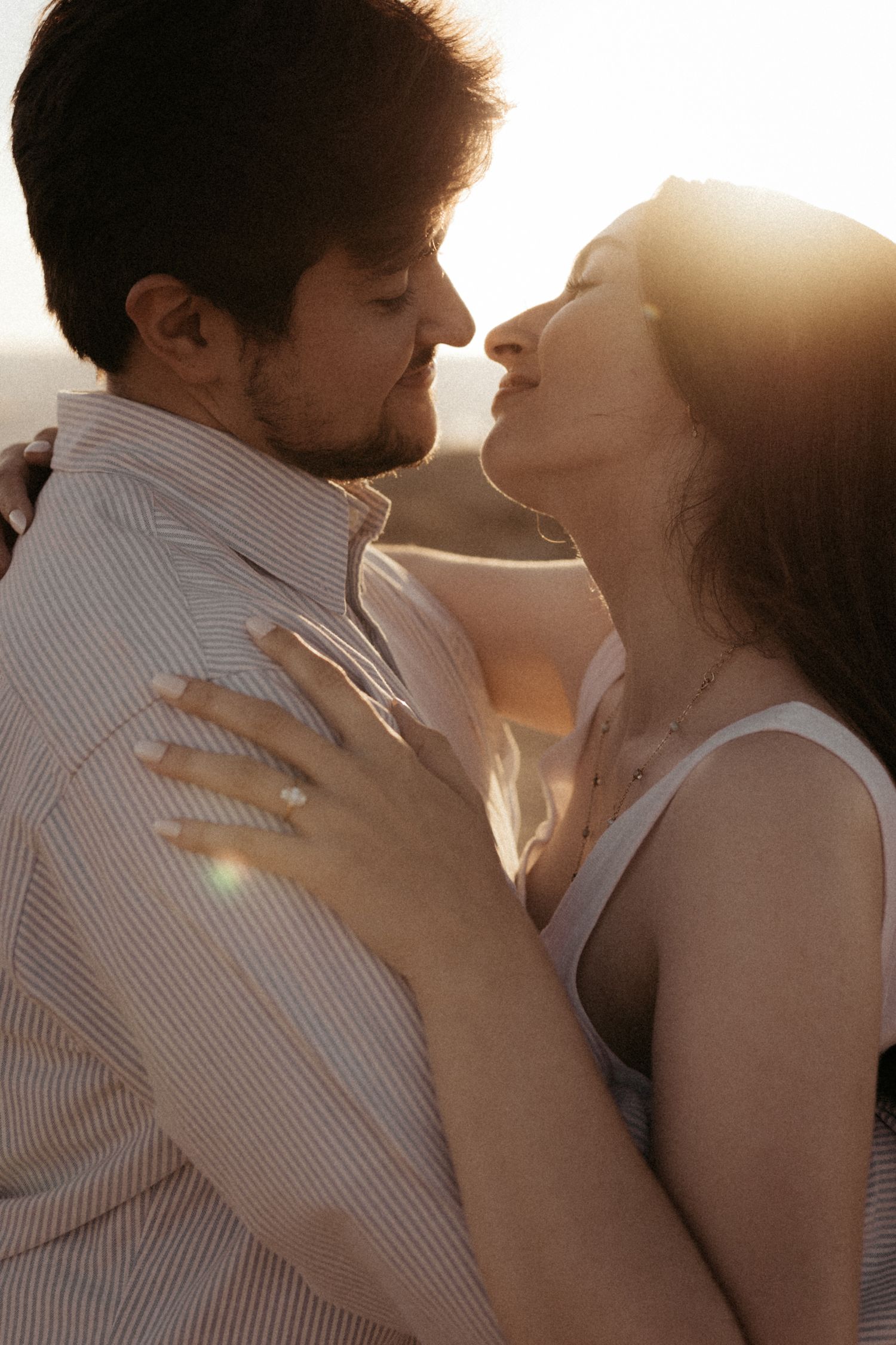 A couple shares a romantic kiss during golden hour with sun flares in the background.