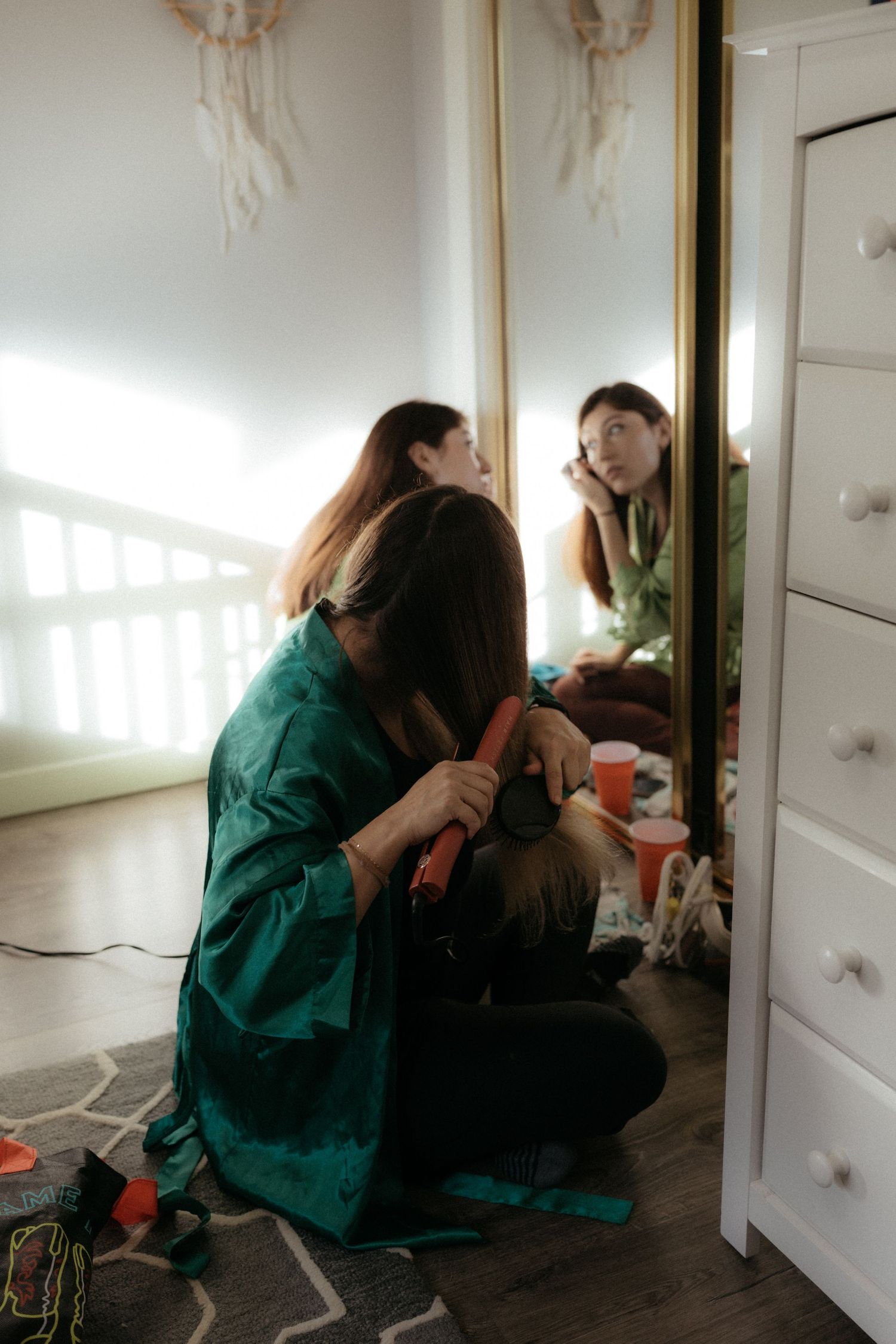 Wedding preparation scene with people getting ready in a bright room with a white dresser and dreamcatchers on the wall.