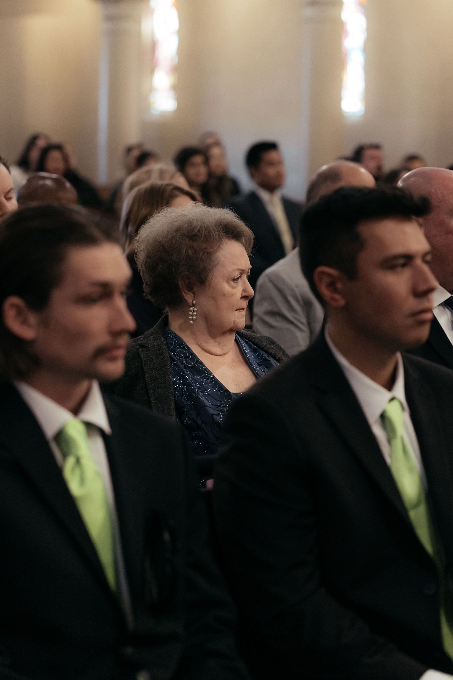 Wedding guests in formal attire seated in church pews with decorative lighting overhead.