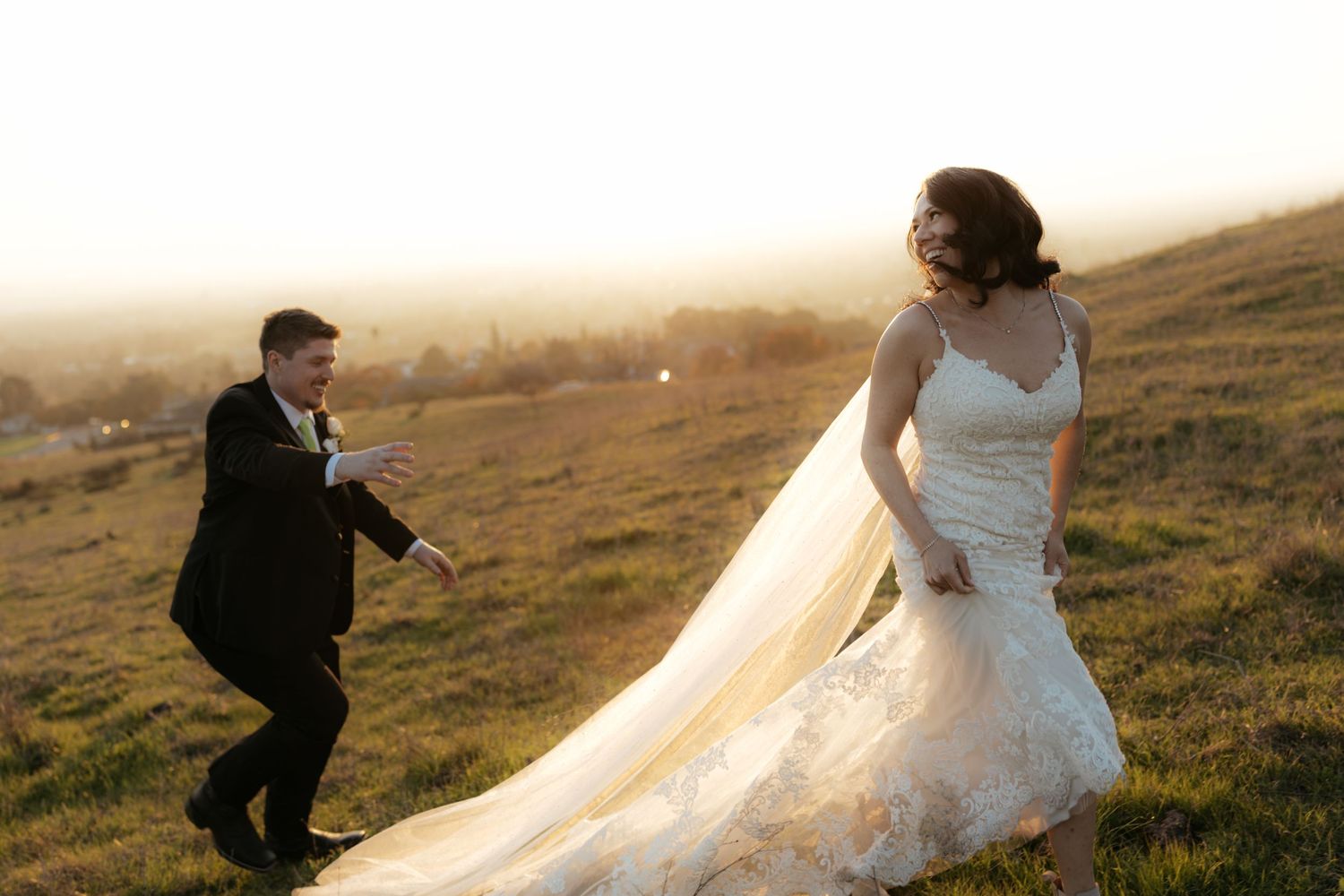 A couple in wedding attire walks along a scenic hillside path during golden hour sunset.