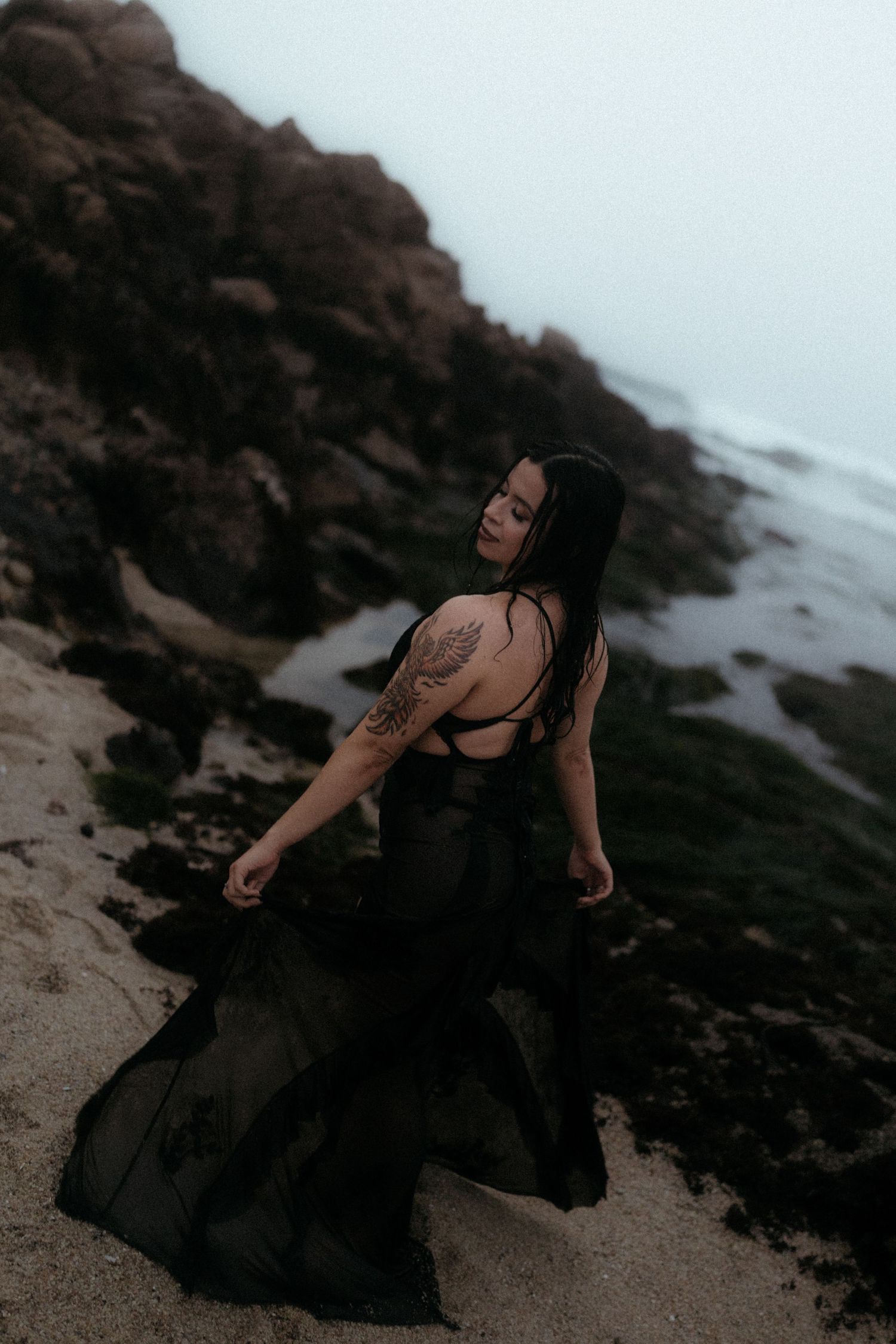 Ethereal beach photoshoot featuring flowing black dress against rocky coastline.