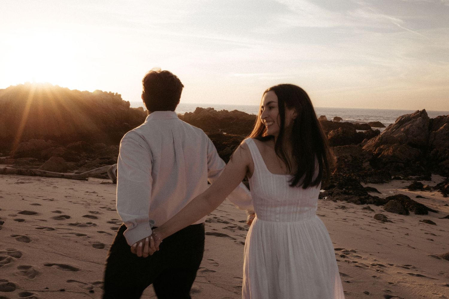 Silhouetted figures embrace on a beach during golden hour with sun rays peeking through.